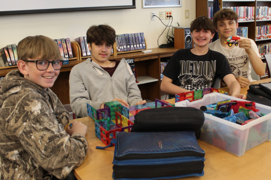 Four boys sit at a table in a library, surrounded by toys and books. They are smiling.