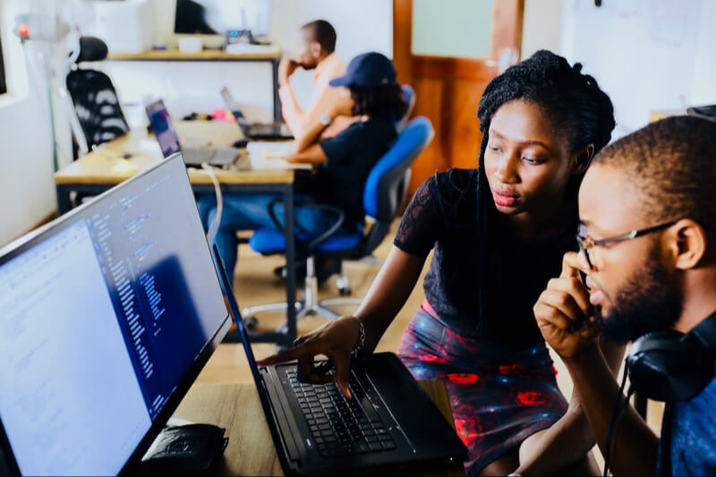 People in an office with desks. Two people focus on a laptop while another person works at a desk.