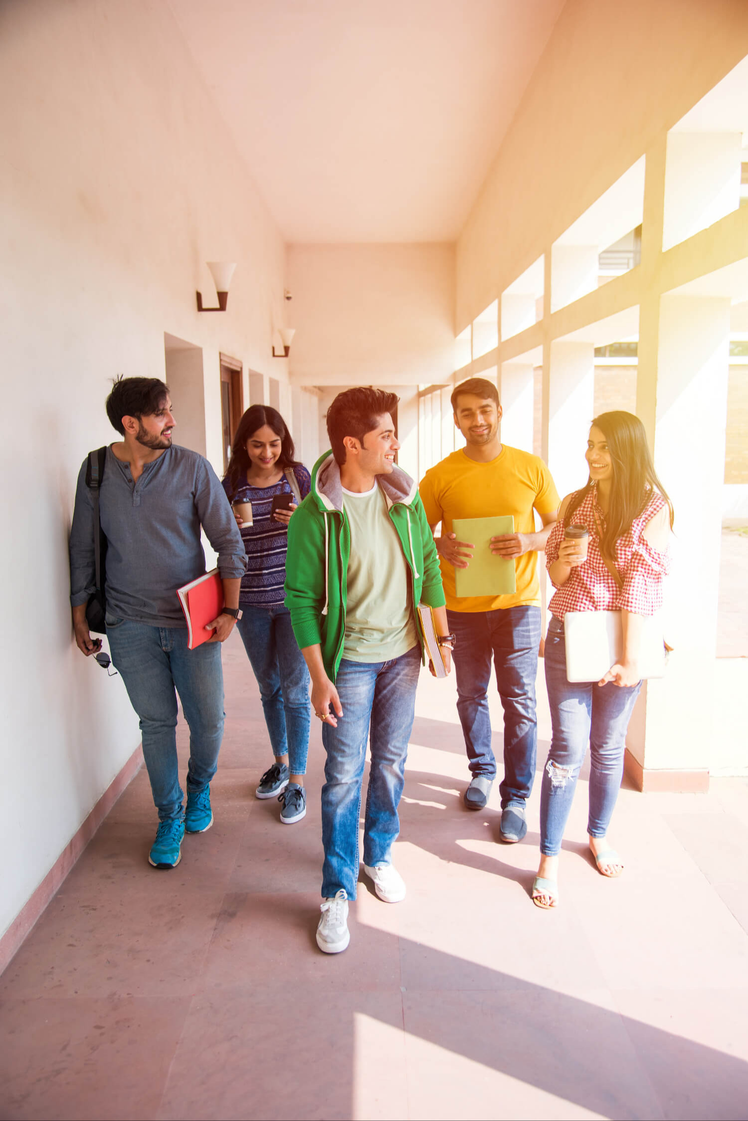 Five individuals walking down a hallway. They hold books and appear to be conversing. Walls and windows line the hallway.