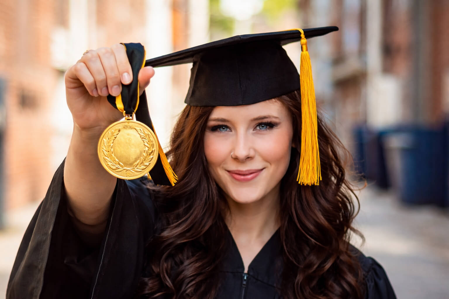 A woman in a black graduation gown and hat holds a gold medal. She smiles at the camera.