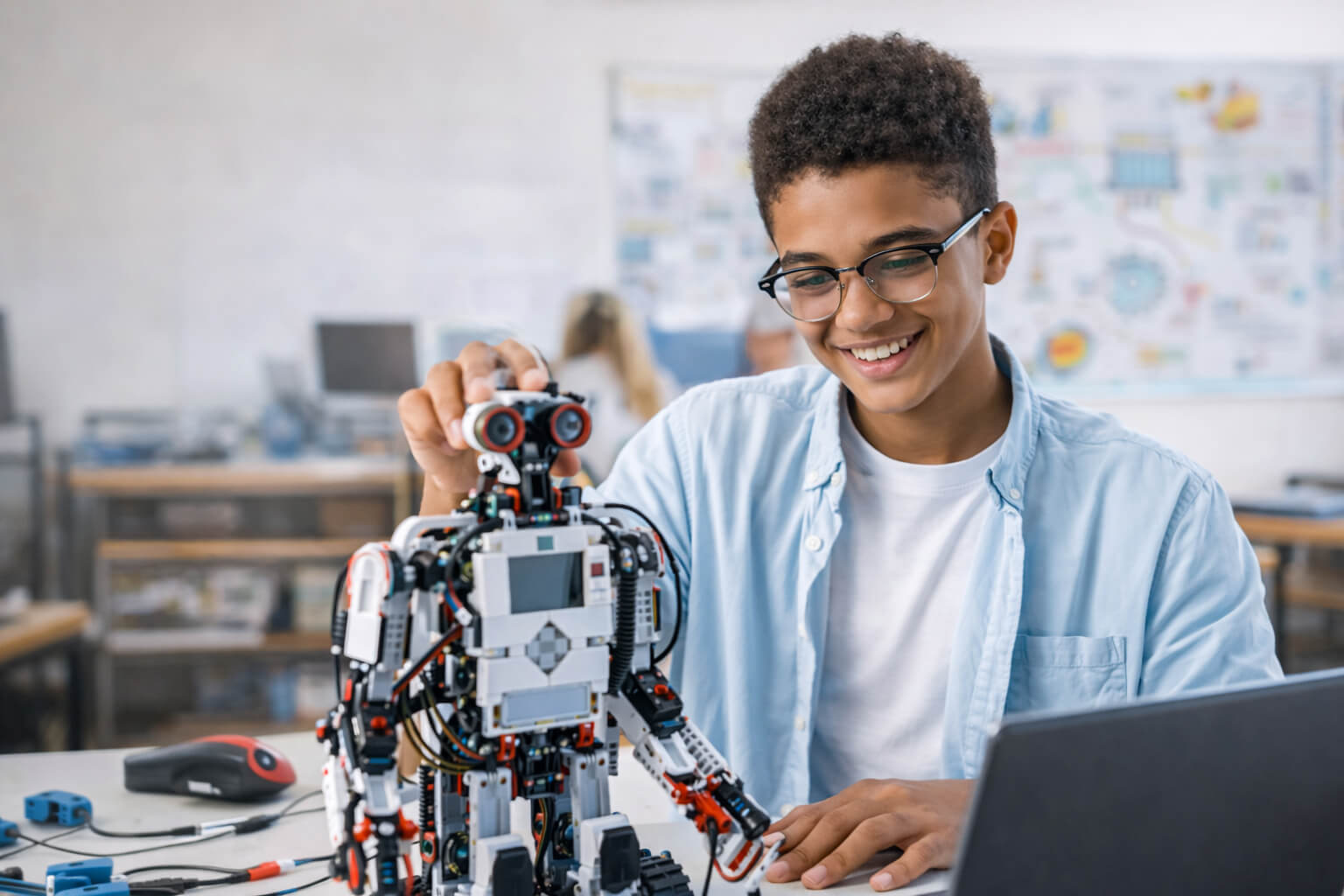 A young person wearing glasses and a blue shirt works on a robotic model next to an open laptop.