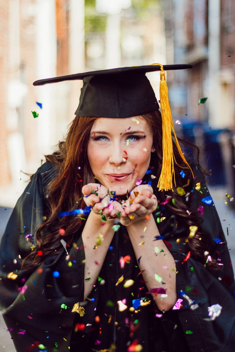Woman in cap and gown with tassel, smiling, surrounded by colorful confetti, on an urban street.
