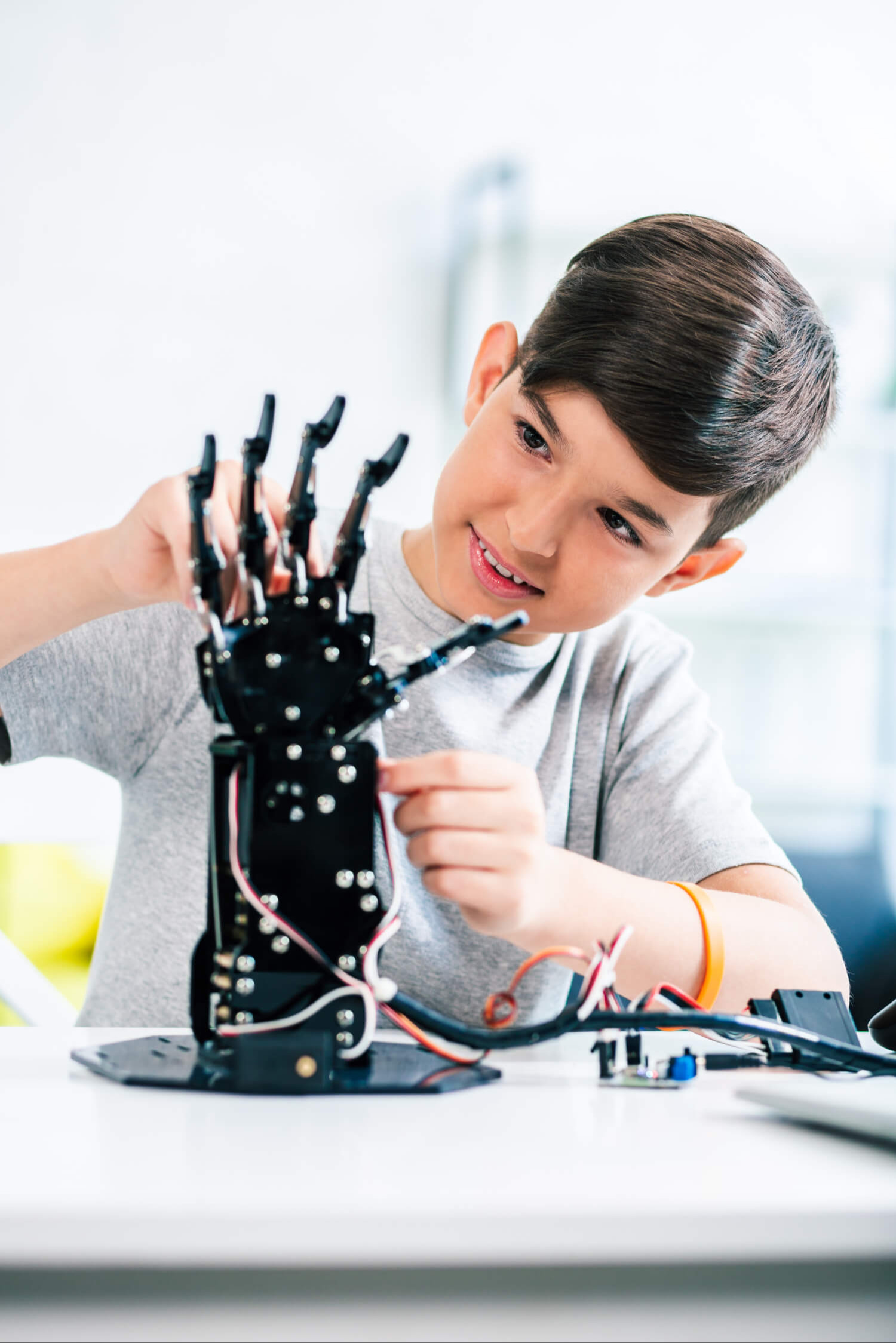 A young boy wearing a gray t-shirt and a bracelet is working on a mechanical hand with wires.