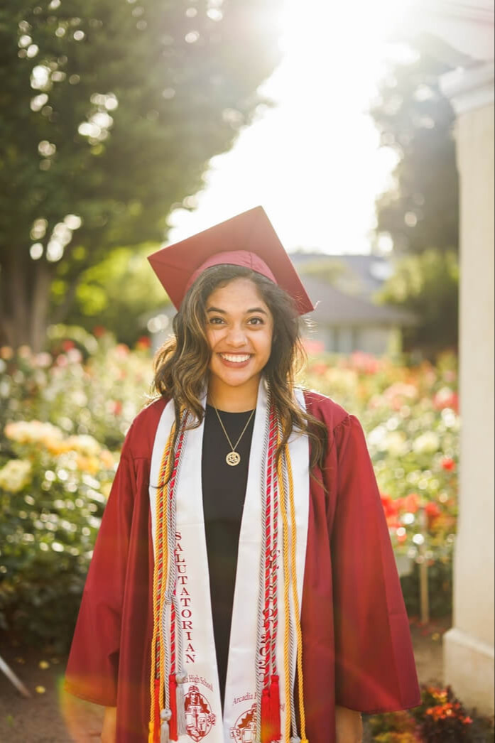 A smiling individual in a red graduation gown, cap, and stole stands in a garden with vibrant flowers.