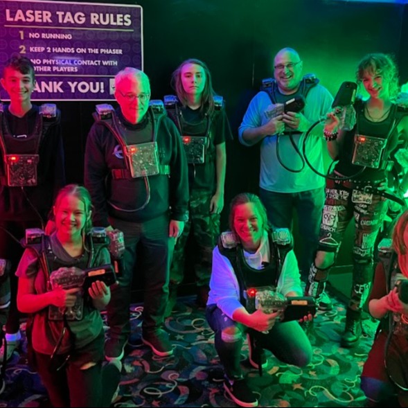 students and adults dressed in laser tag gear with a sign in background that says Laser tag rules, Everyone is lit with a neon colored lighting and is smiling at the camera