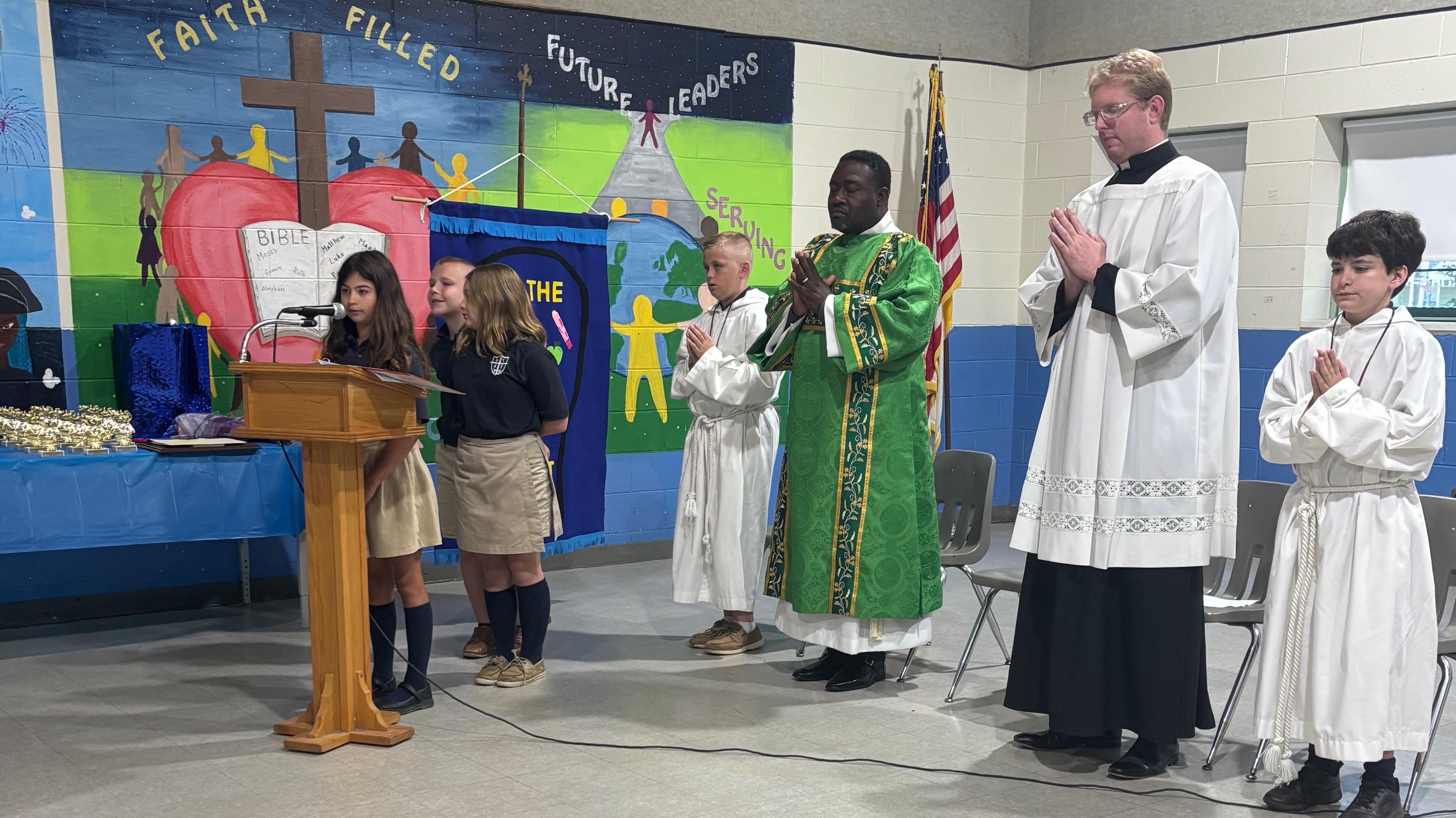 Four children in white robes stand by a podium. One child speaks into a microphone. Behind them, a priest in green and white stands at a podium, and a flag stands nearby.