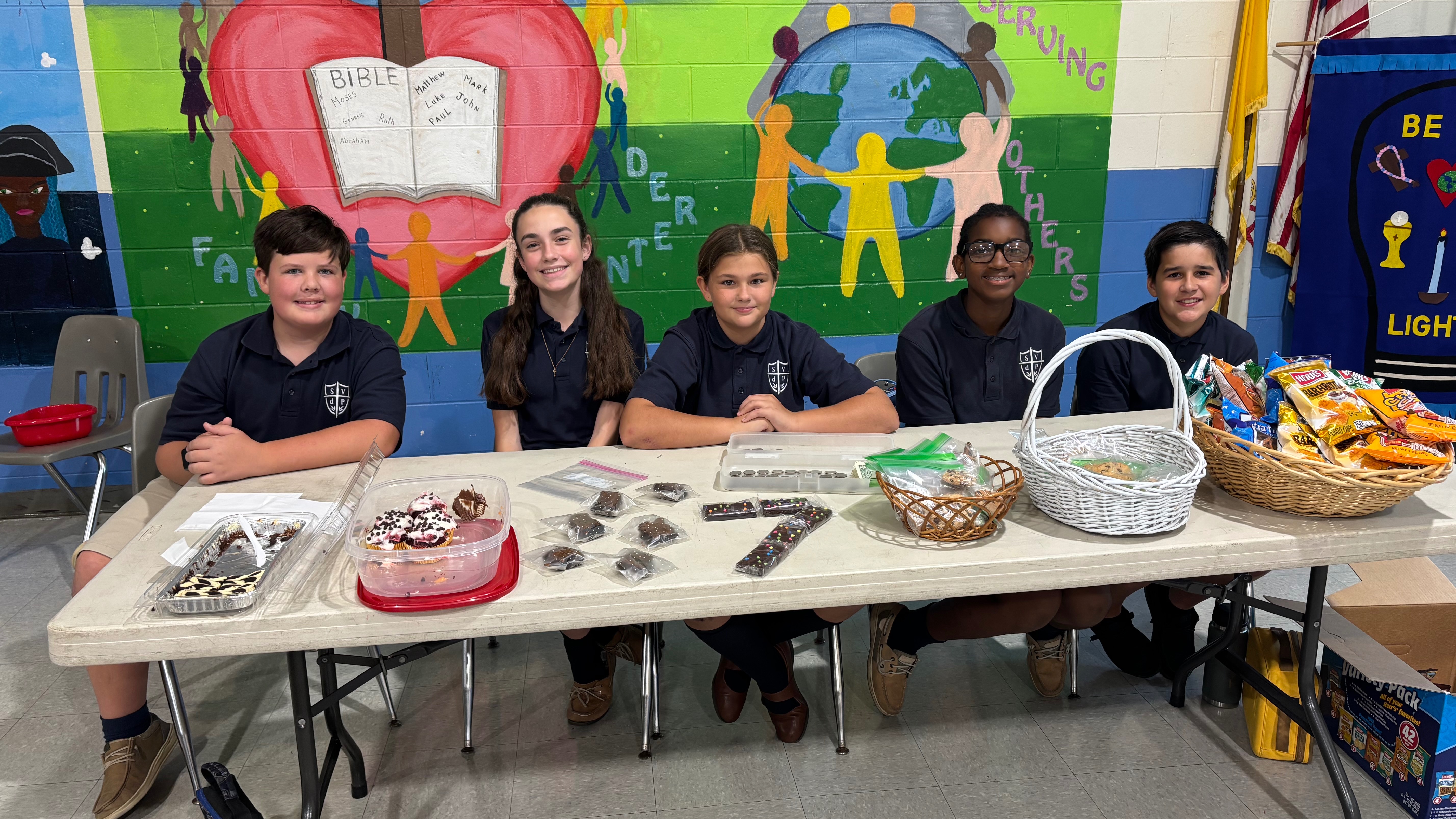Five students sit at a table with cookies, a cake, and baskets. A mural with various symbols is behind them.