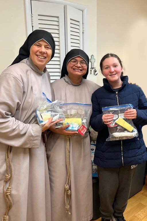 Four women in robes, one wearing a blue jacket, holding packages with food, standing in a room with wood floors.