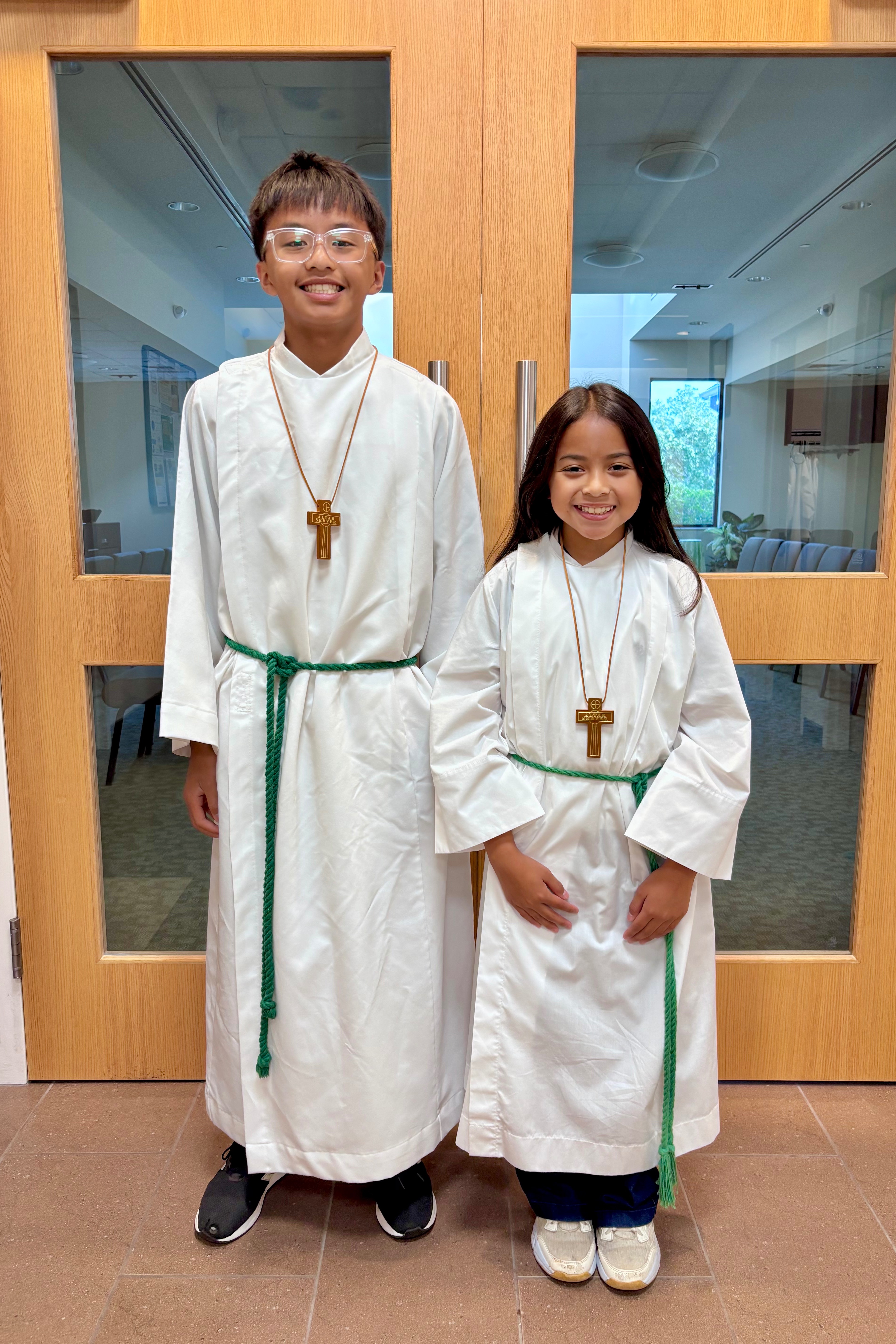 Two children in white robes and green sashes with visible crosses around their necks, standing next to each other in front of a wooden door.