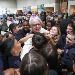 Many children and a woman, wearing glasses, are smiling in a classroom. The woman is surrounded by children.