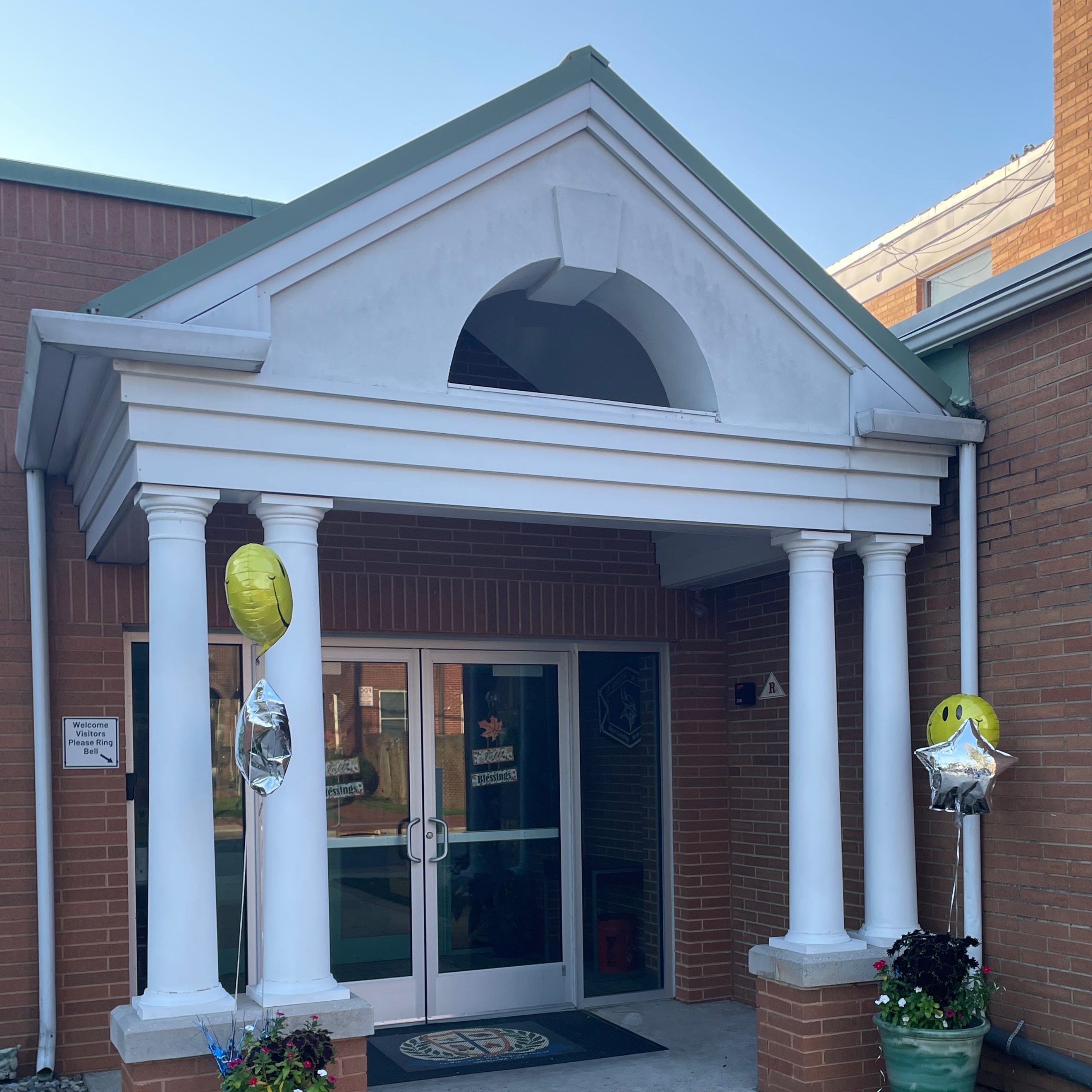 A building with a white archway entrance, pillars, and two green potted plants. Balloons are on either side.