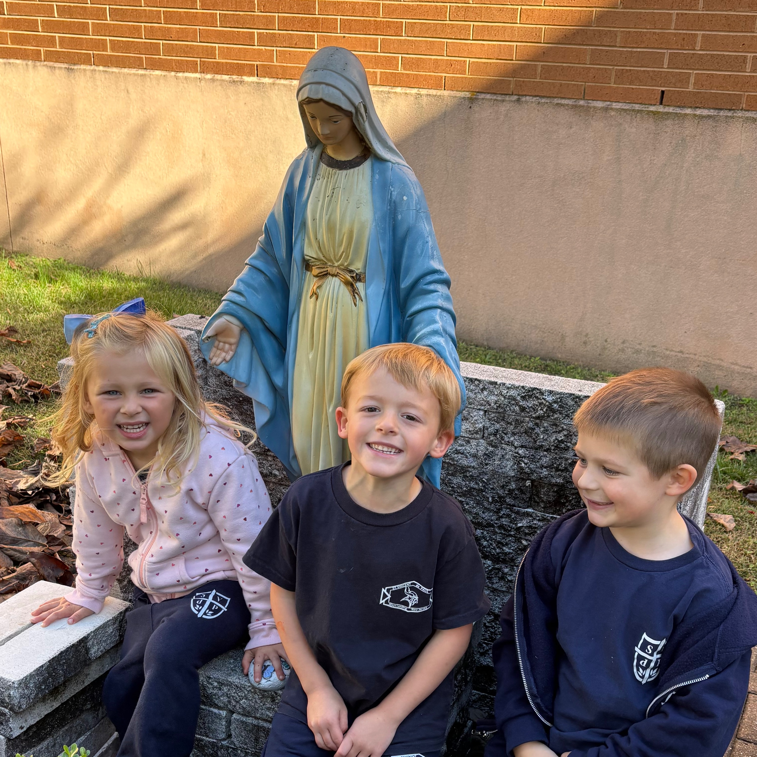 Four children sit in front of a statue of a woman, on a stone bench. Behind them is a brick wall.