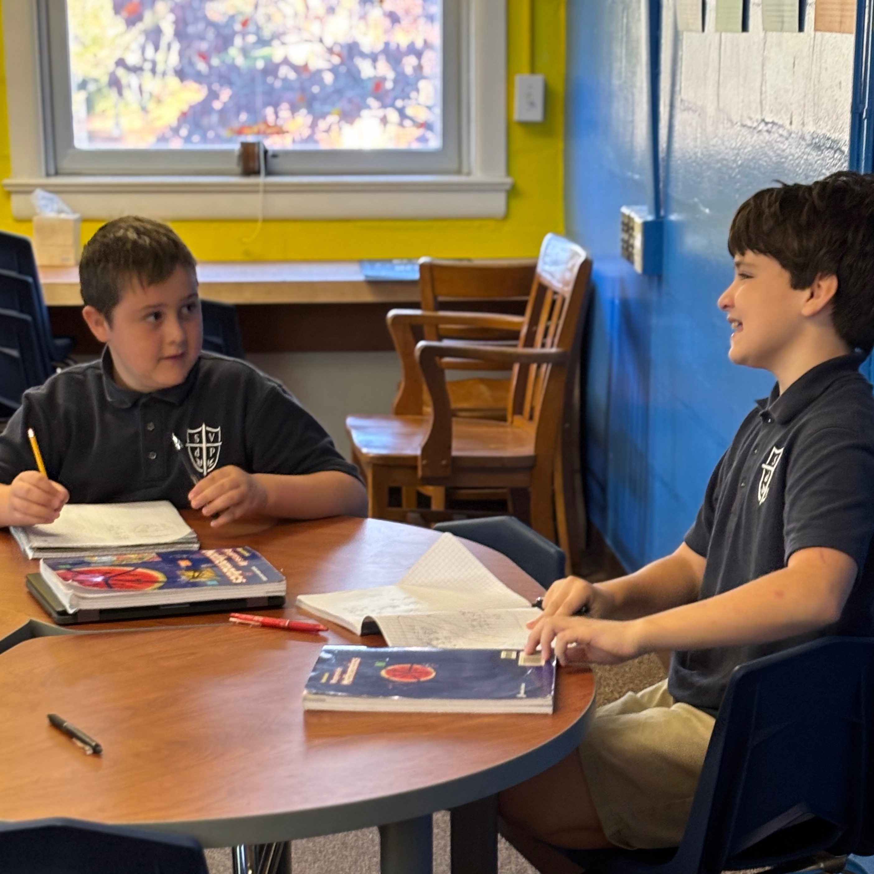 Two boys in a classroom. One boy holds a pen and the other boy holds a book.