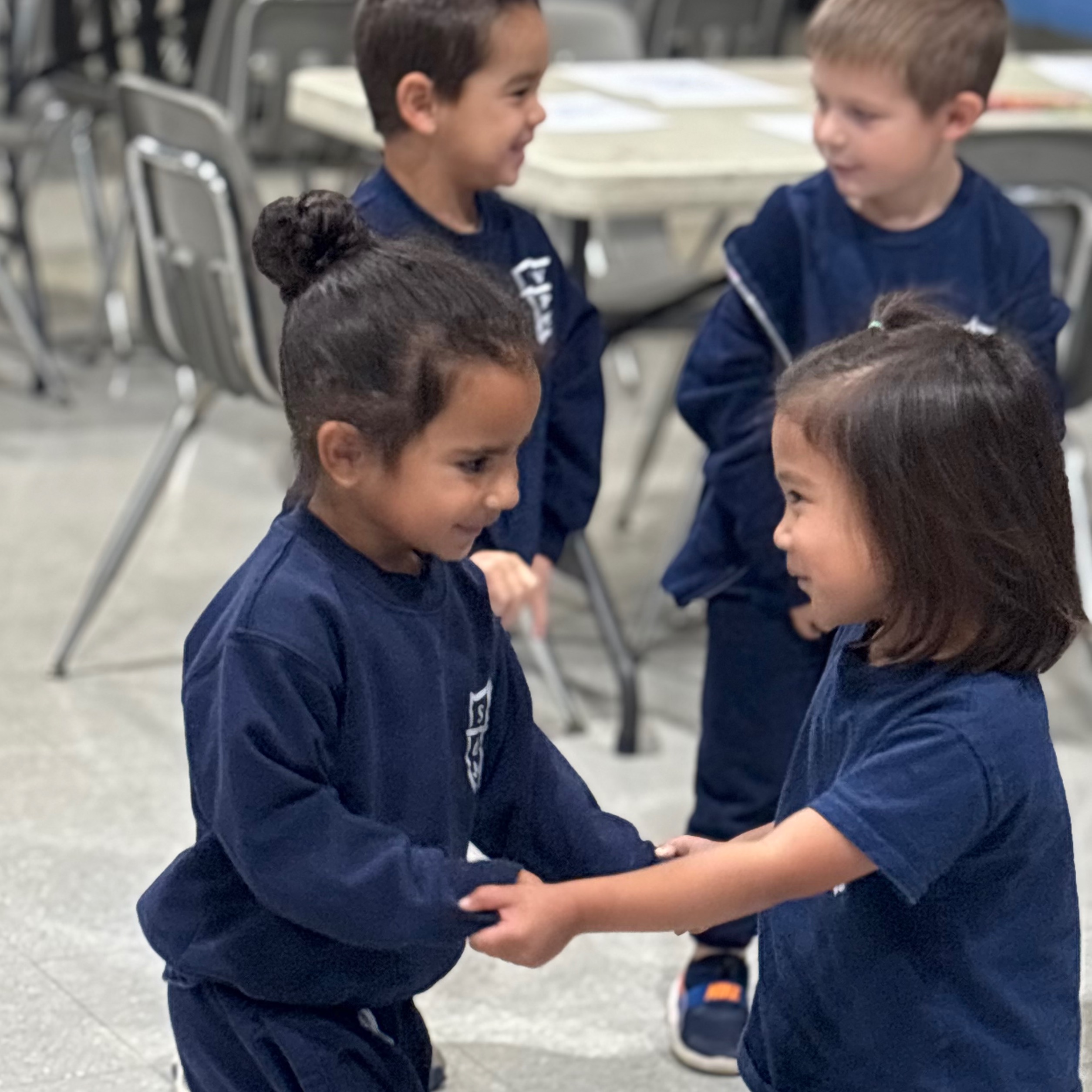 Four young children wearing matching navy blue uniforms interact in a room. Two girls hold hands.