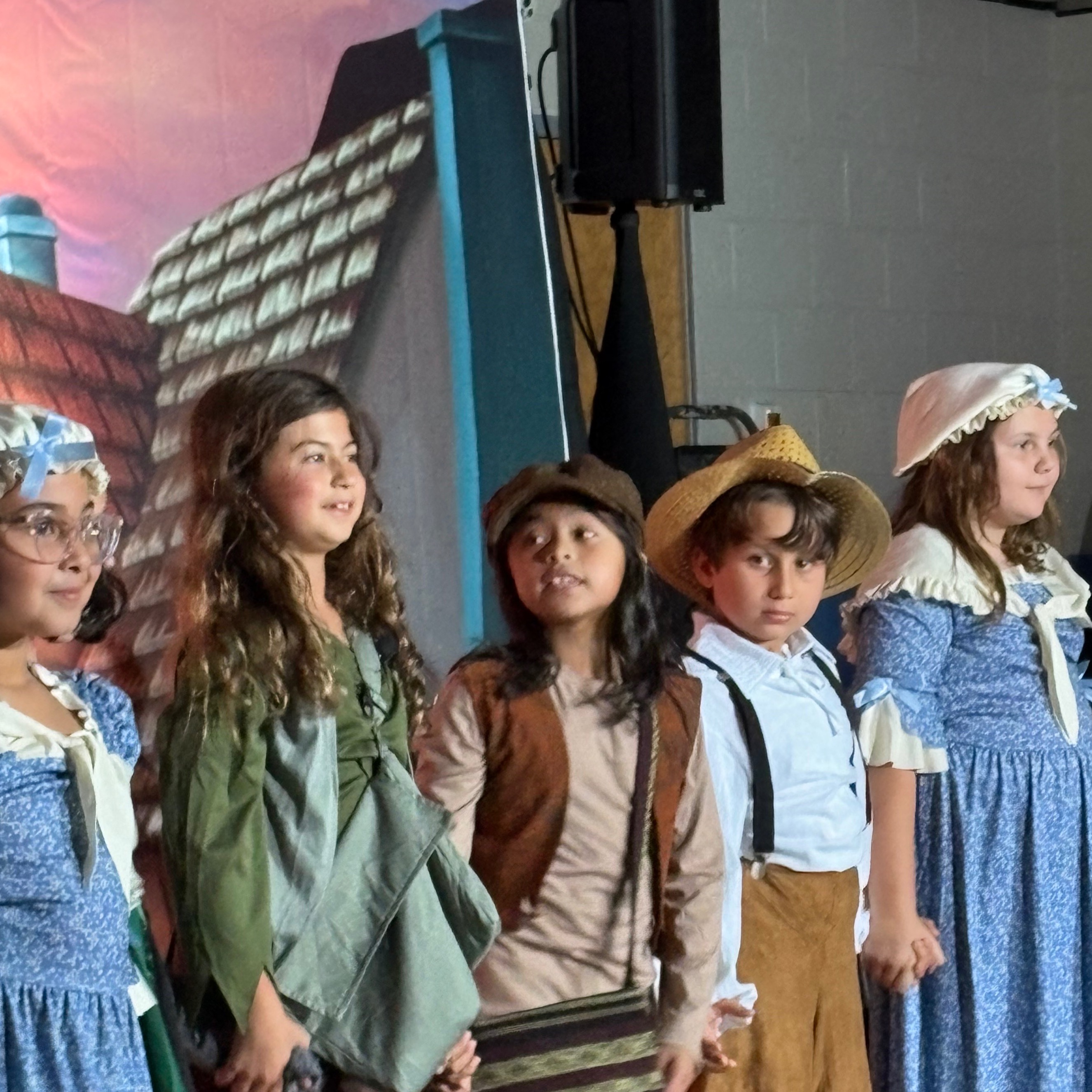 Kids in historical costumes stand in a row on a stage with a painted background.