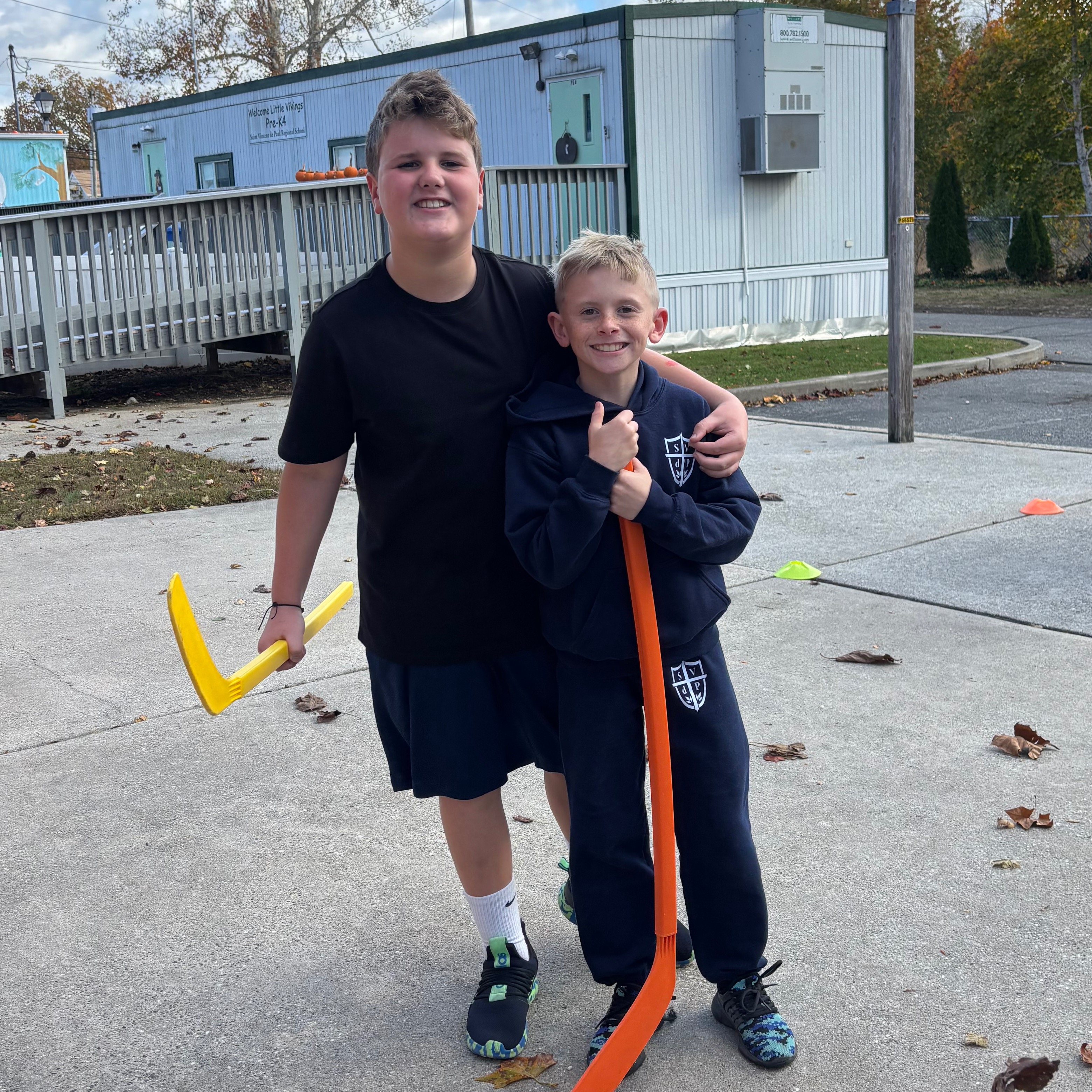 Two children holding hockey sticks in an outdoor area with a building and trees in the background.