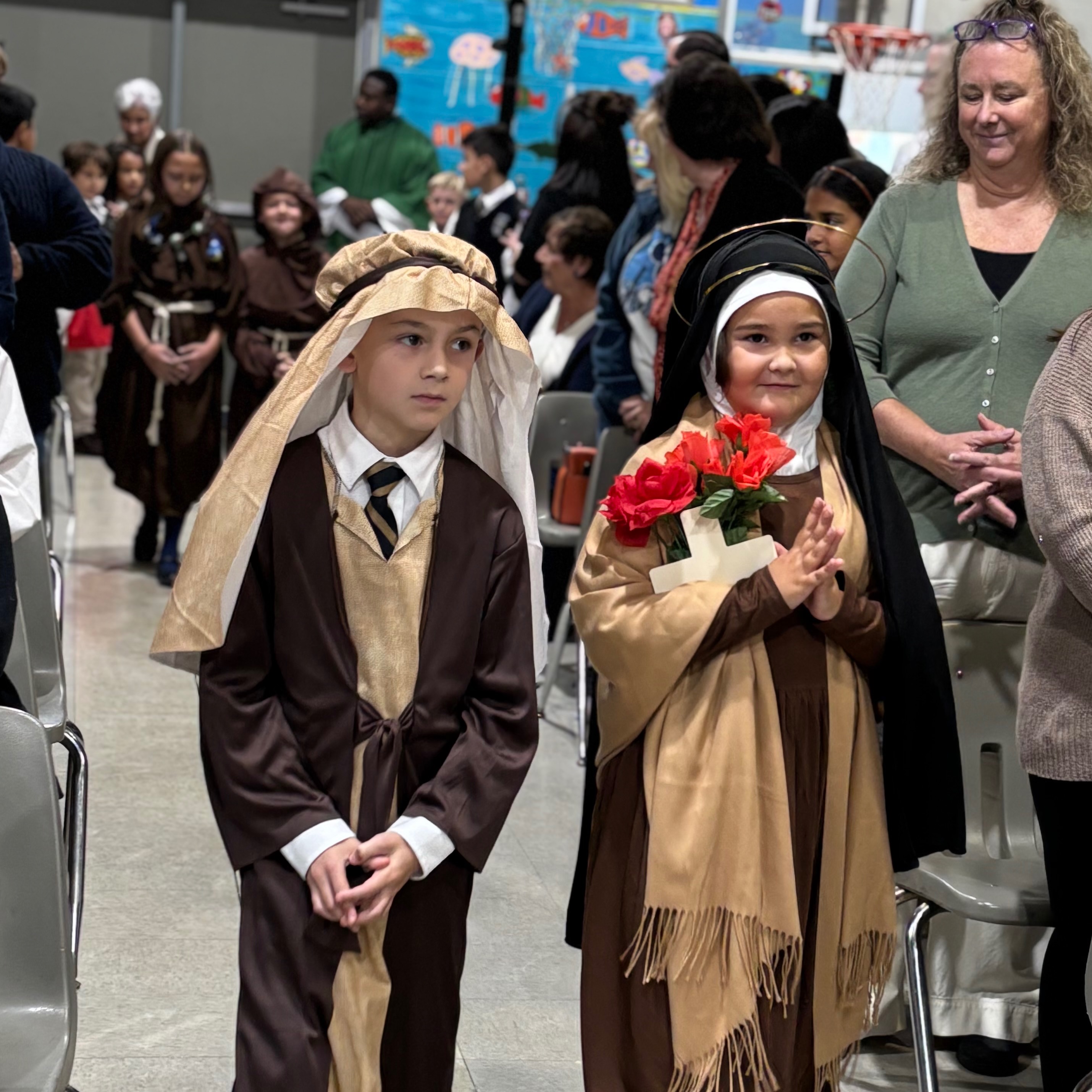 Children dressed in costumes, including a nun's habit, walk down a row of chairs. Some adults stand behind them.