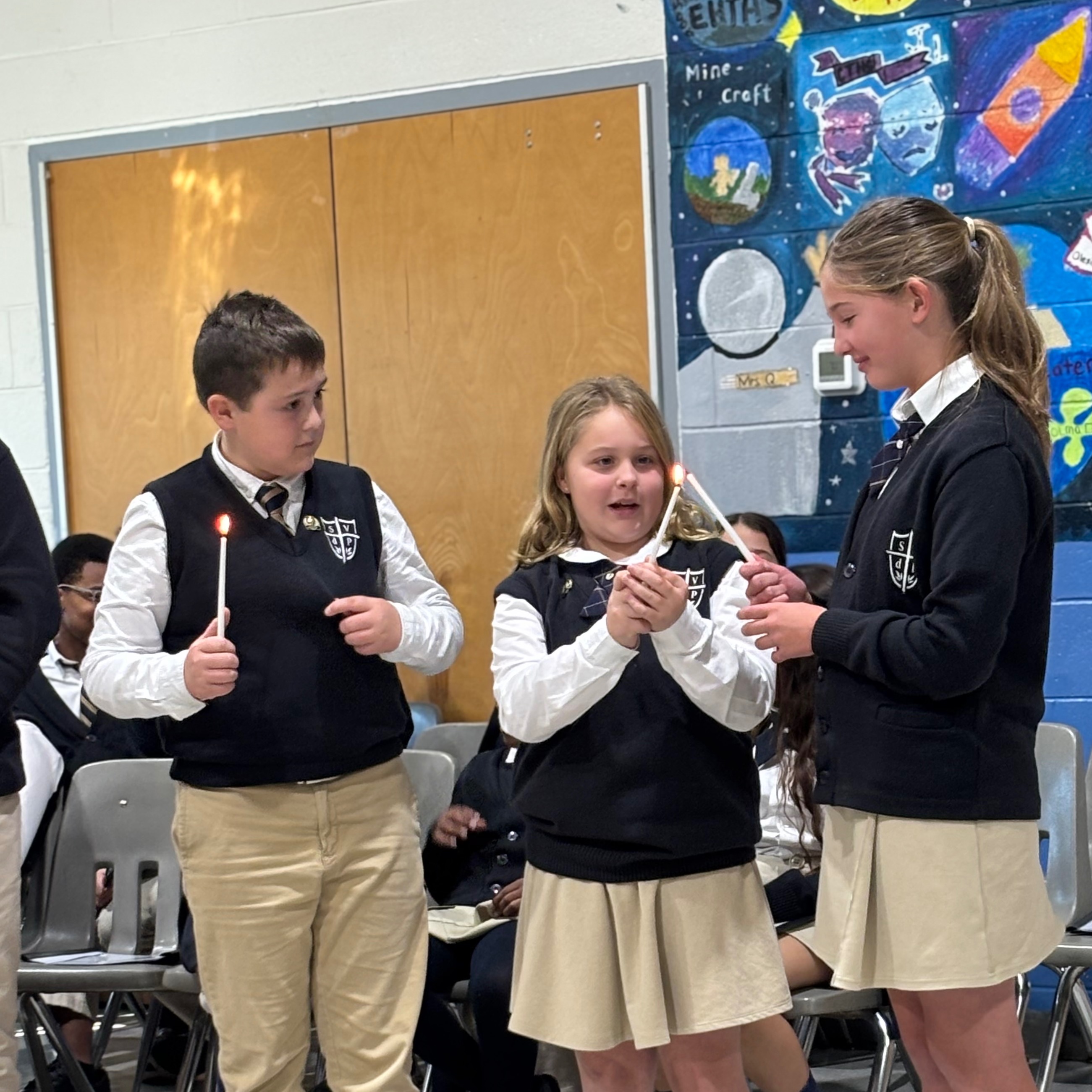 Three students stand holding candles in a classroom. They wear matching uniforms and shoes. A person sits in a chair behind them.