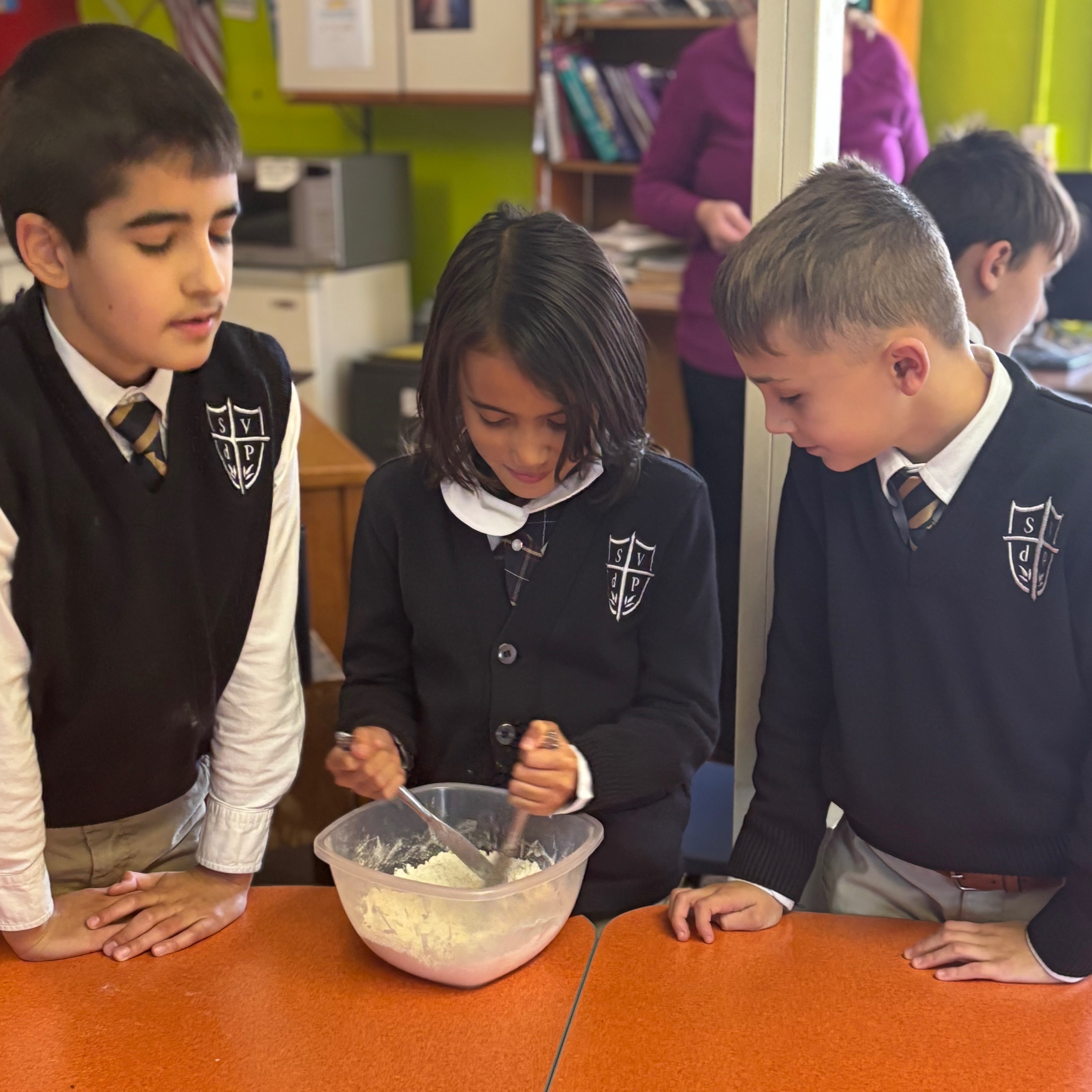 Three children in uniforms over a table, one with a rolling pin, another with a bowl of dough.