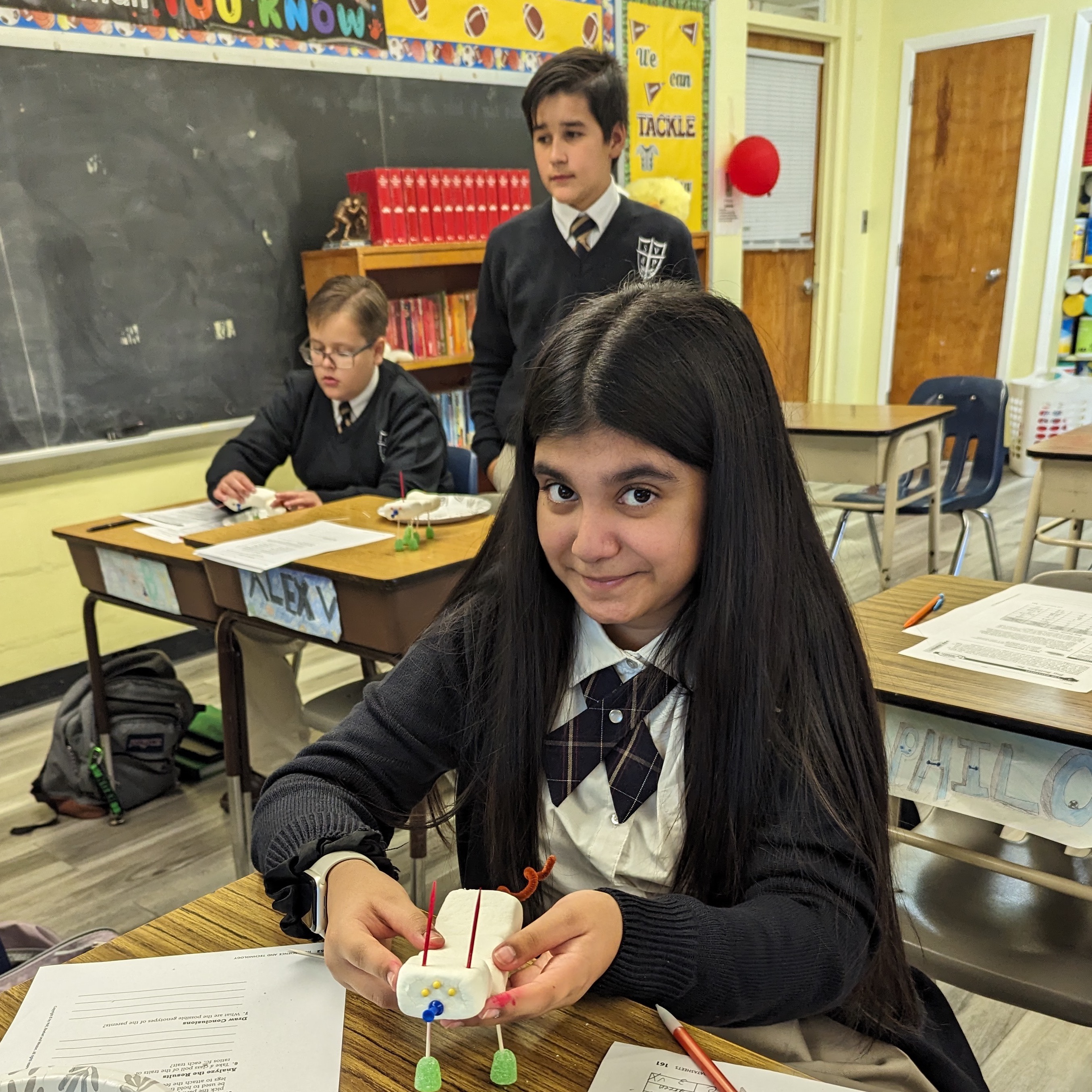 Students in a classroom with wooden desks and chairs, engaged in activities. A girl is holding a small object, and there is a blue binder on a desk.