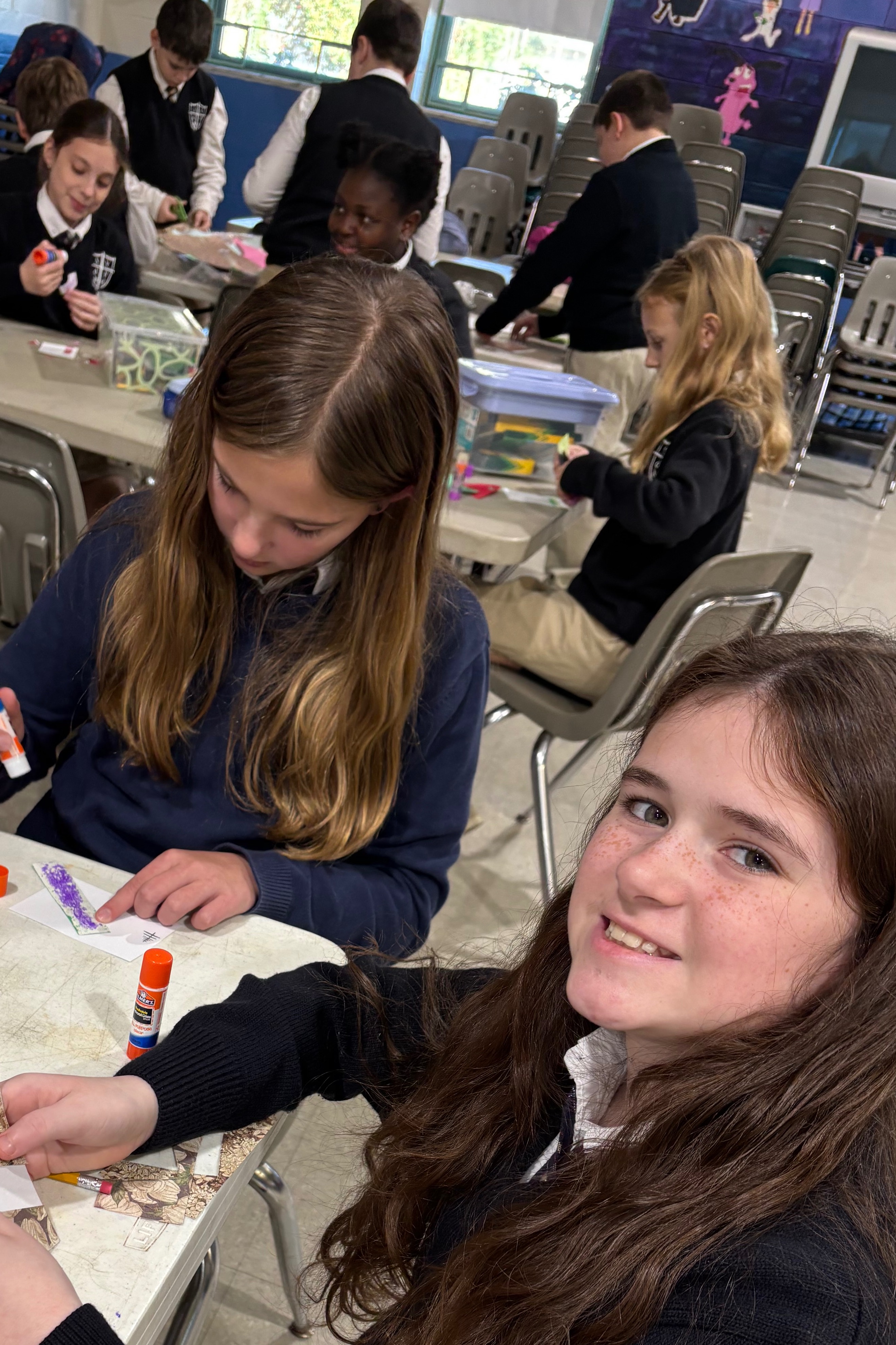 Students sitting at tables, doing crafts. A girl in a black shirt smiles, with a red bag nearby.