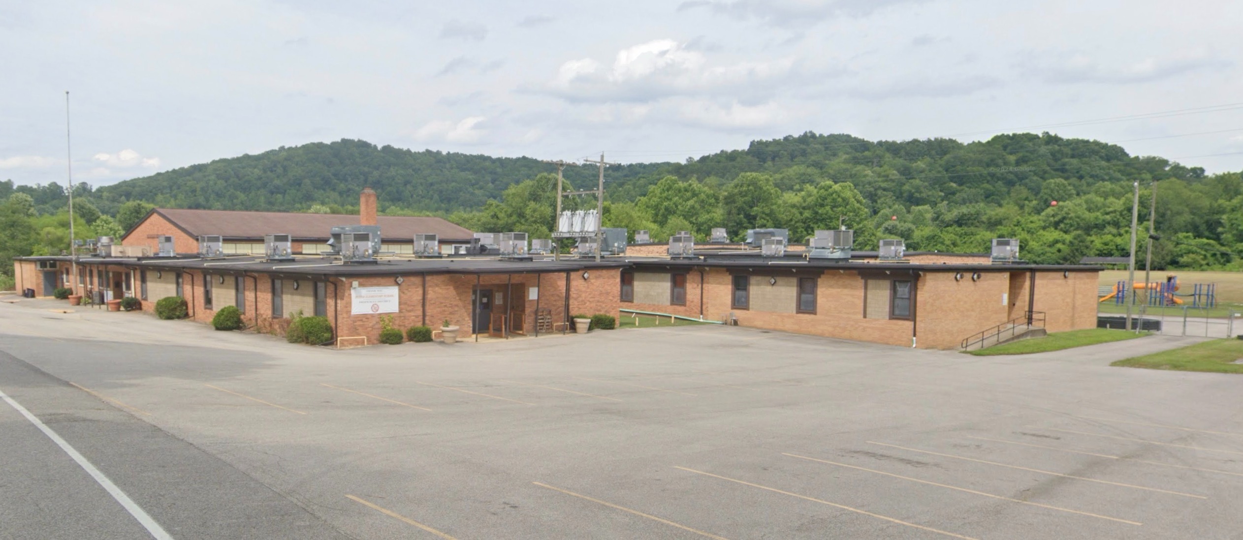 Large building with solar panels on roof, set back from road; empty parking lot; hills in background.