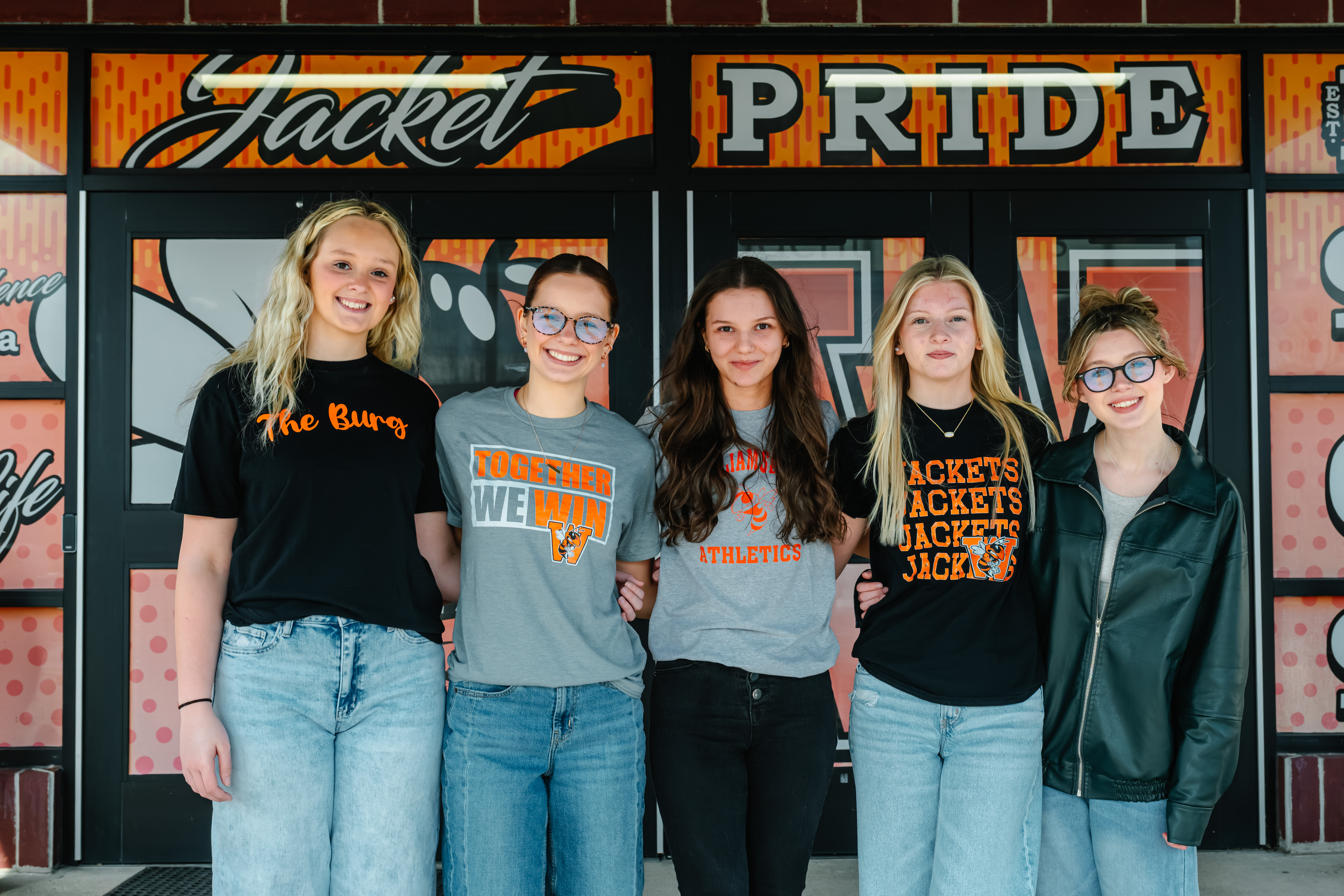 Five women stand before a wall with orange, black, and white writing, including "Jacket Pride." They wear glasses, jackets, and jeans.