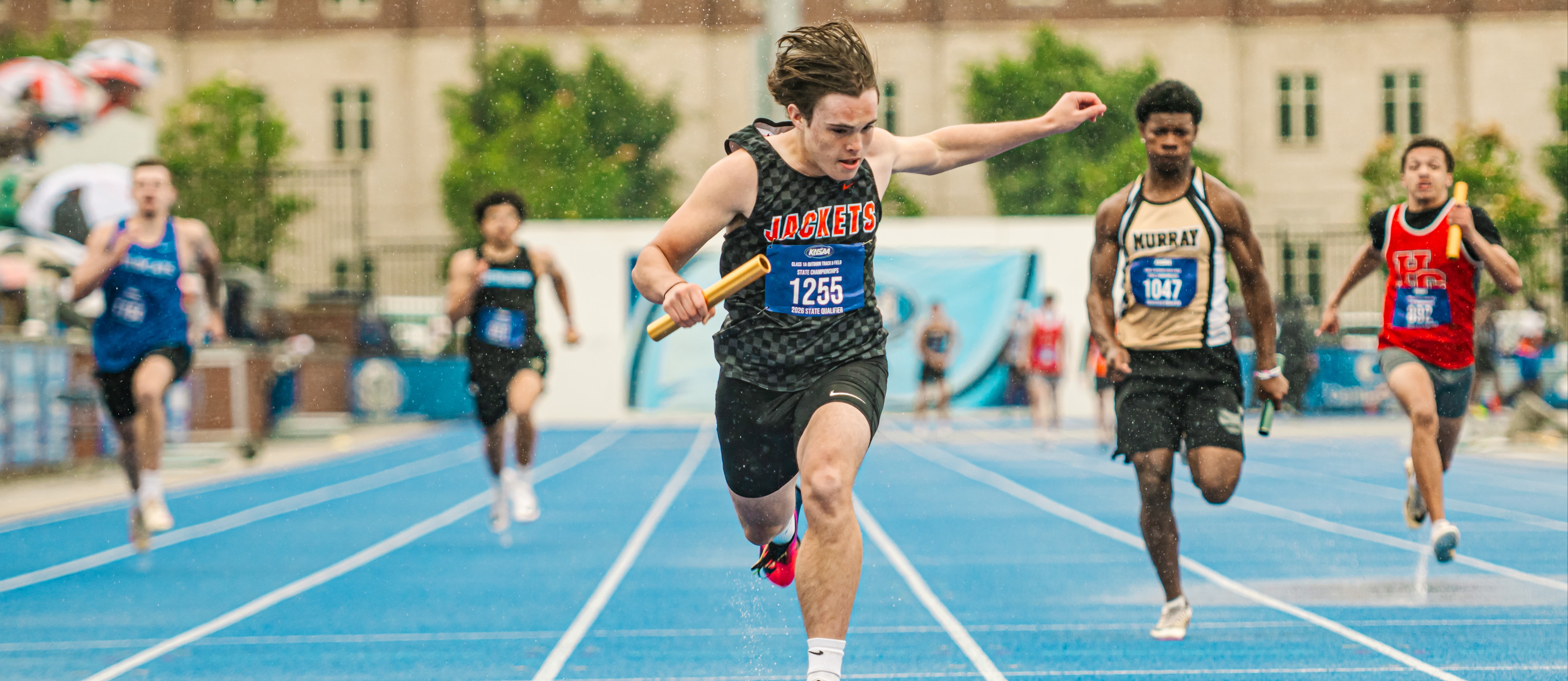 Several athletes sprint on a track with blue lanes; the leading runner holds a baton. Behind them, a building with many windows.