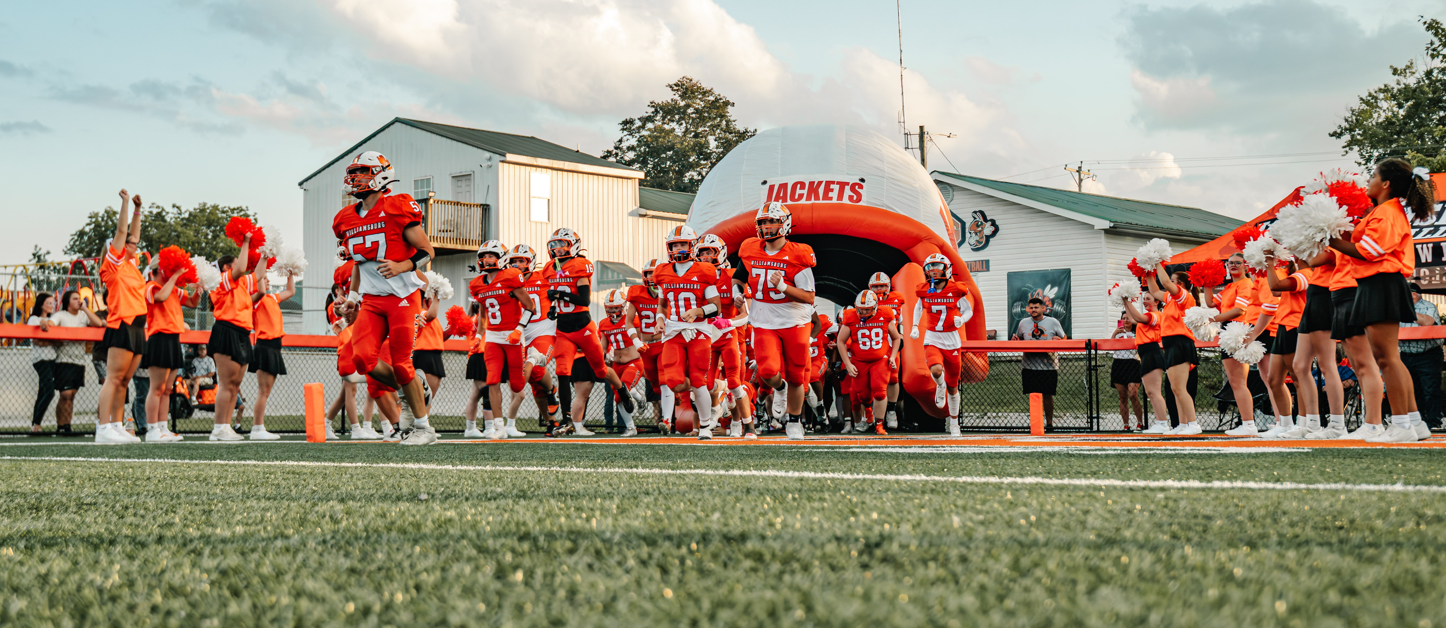 A group of football players in orange uniforms stand on a field. The sky is partly cloudy.