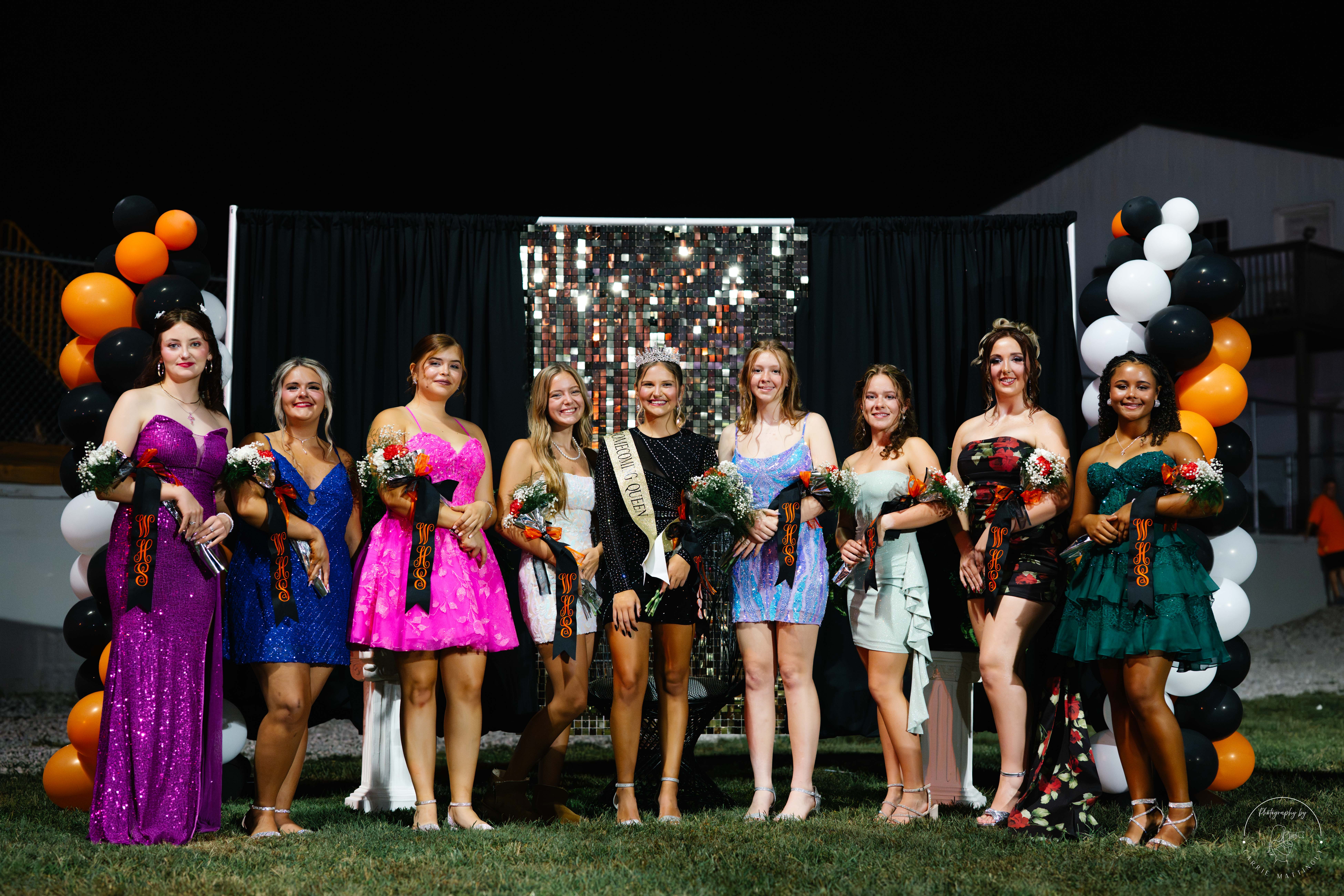 A group of women wearing dresses stand together on grass in front of balloons and a backdrop.