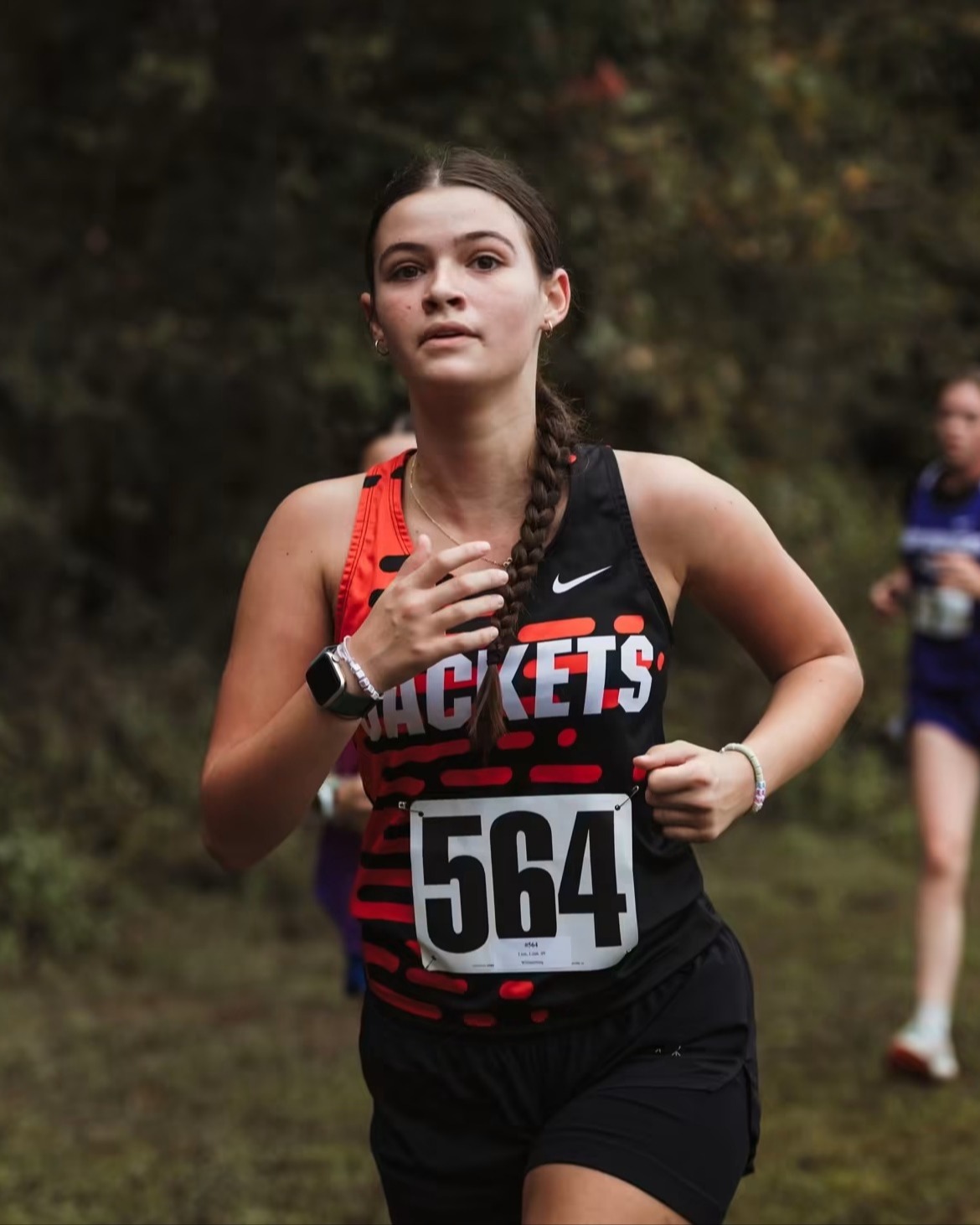 A female runner wearing a black shirt with number 564, followed by two other runners in a grassy field with trees in the background.