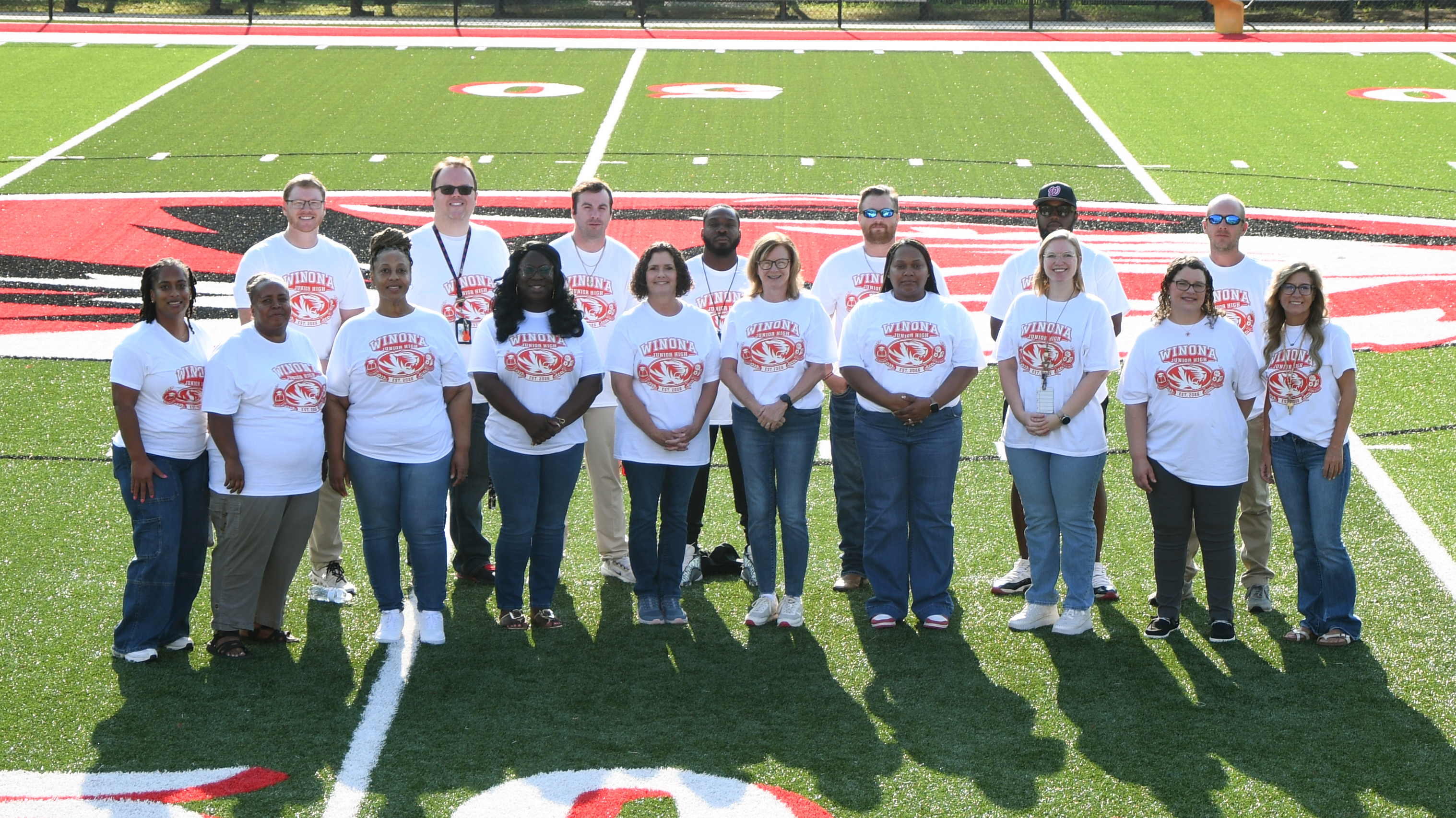 A group of people in white T-shirts with red designs stand in a line on a football field.