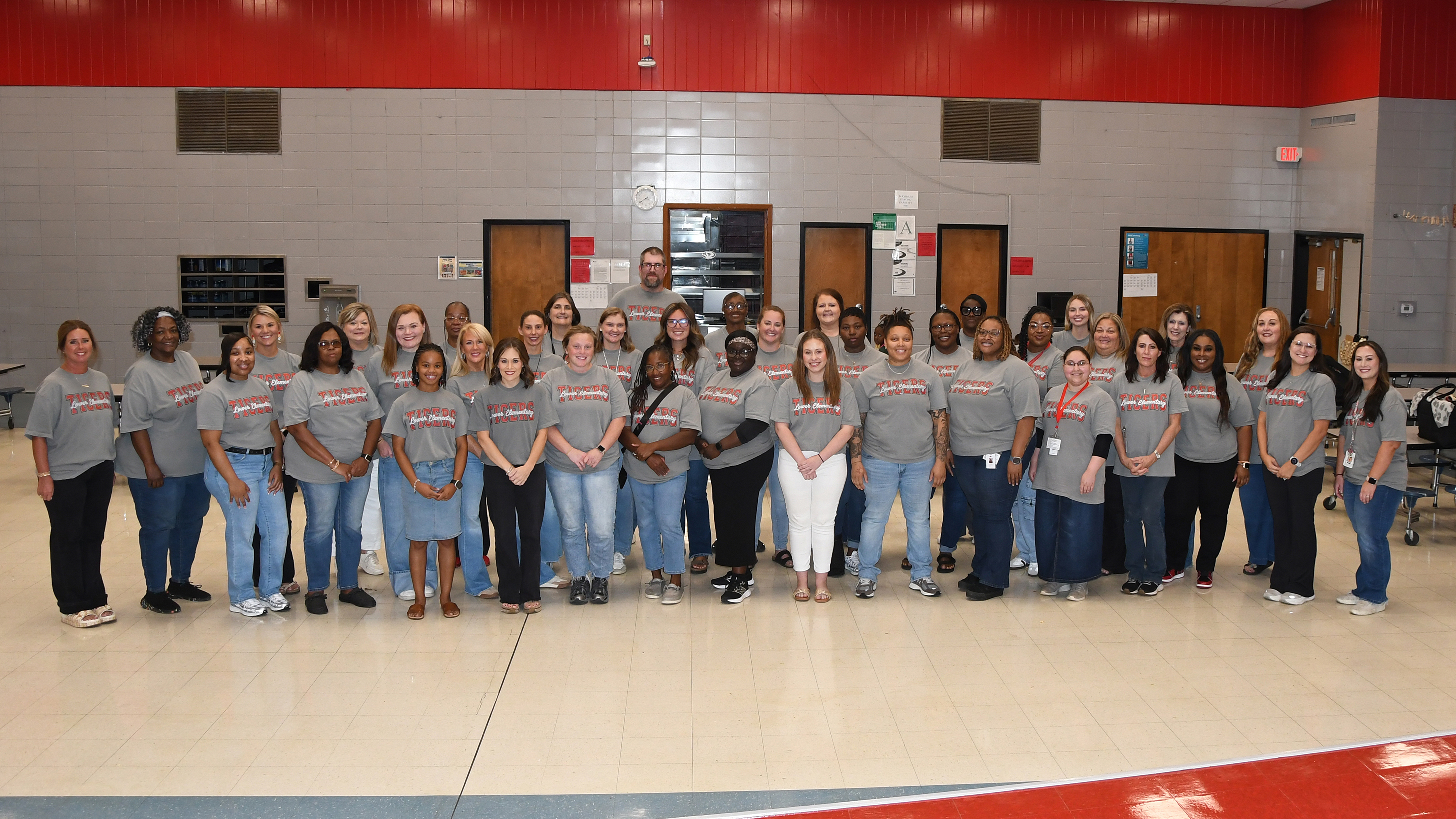 A group of people in gray t-shirts and jeans pose for a photo in a gym with red and white walls.