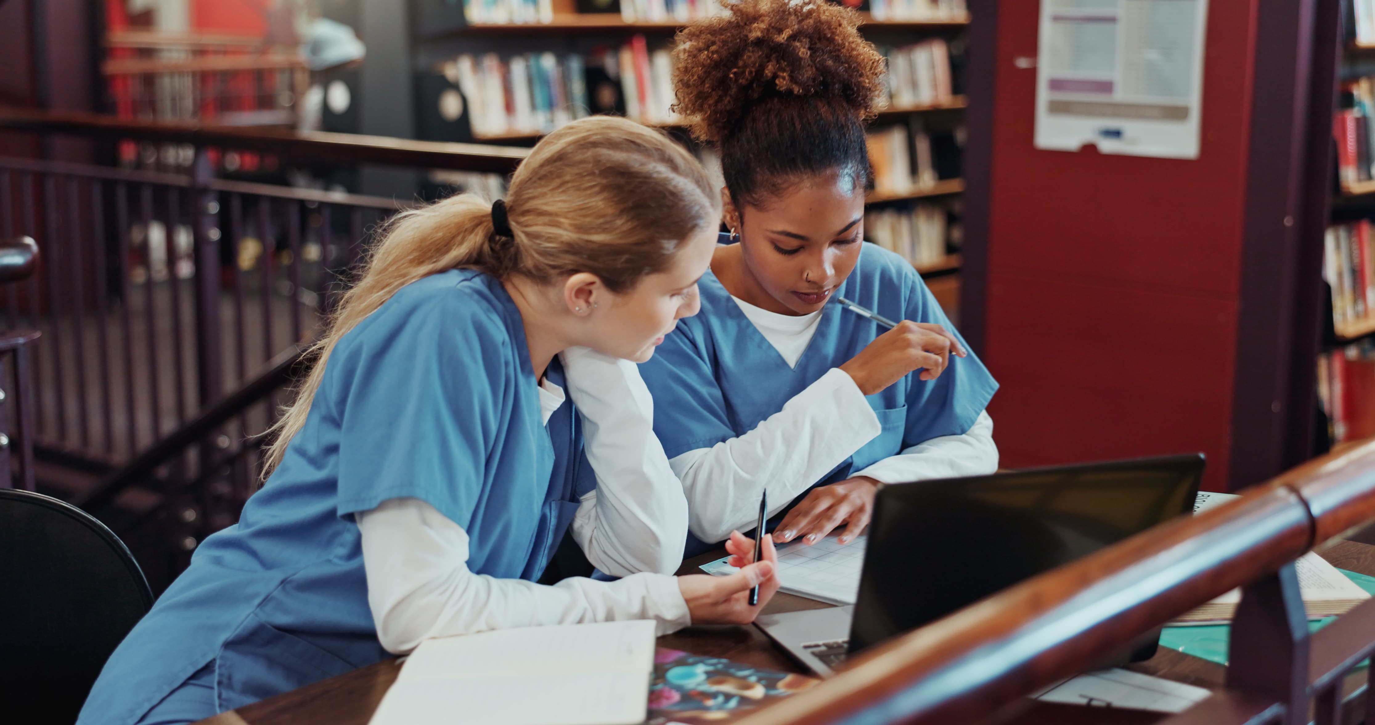 Two women in blue scrubs sit at a table, focusing on a laptop with papers and a pen.