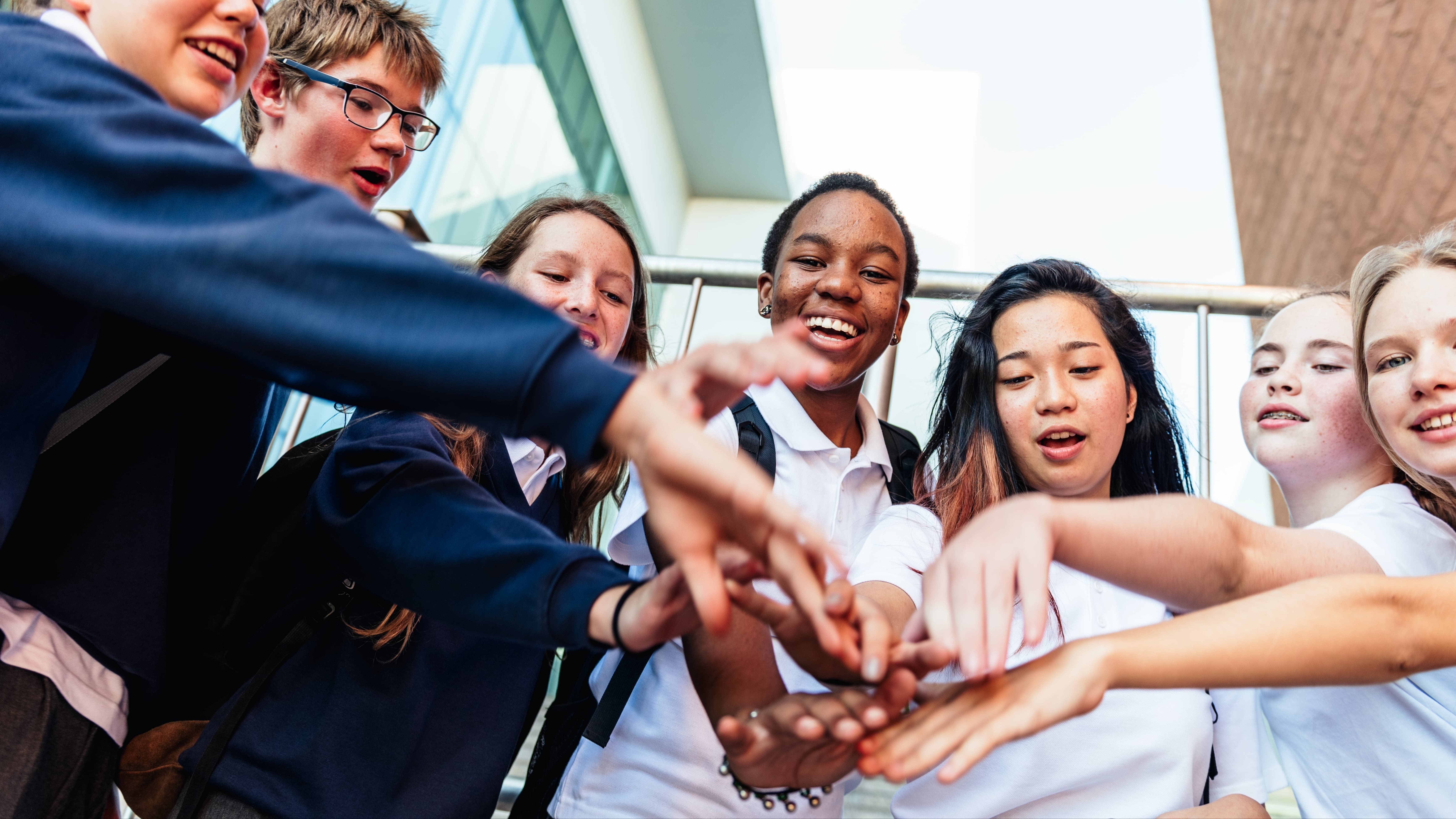 A group of students with hands stacked in a huddle, smiling and looking forward in a bright room.