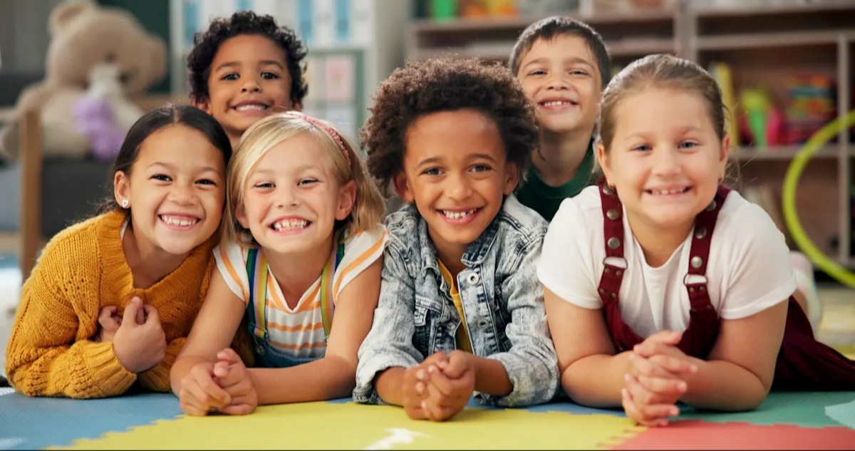 Kids smiling, lying on floor, wearing sweaters and overalls, some holding hands, in a room.