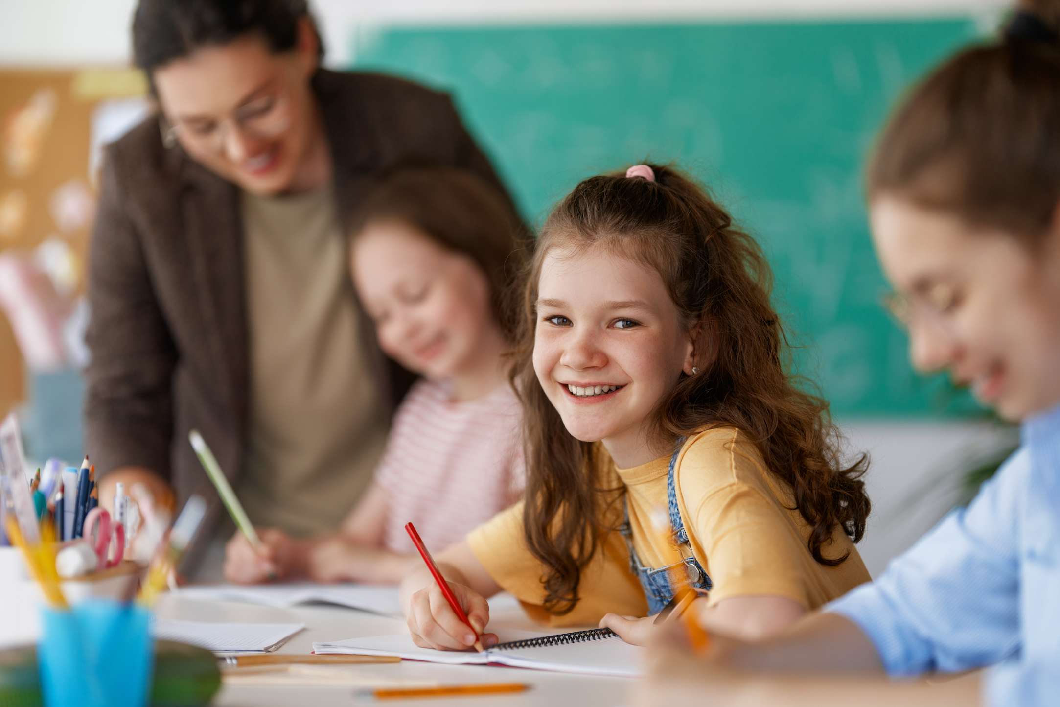 A teacher and three children are seated in a classroom. The children are writing with pencils.