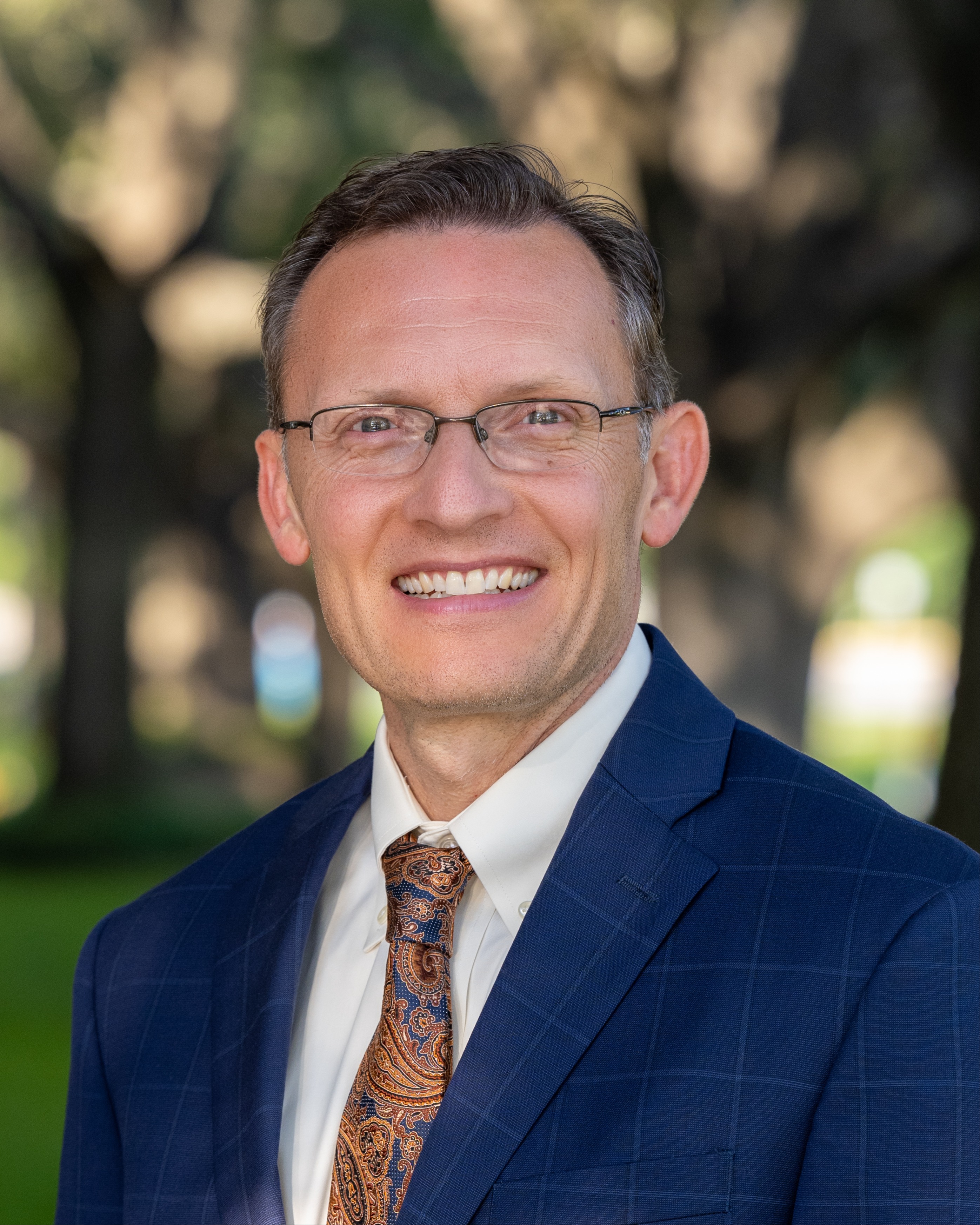 A man in a blue suit, white shirt, and tie smiles. He wears glasses. Trees and grass are in the background.