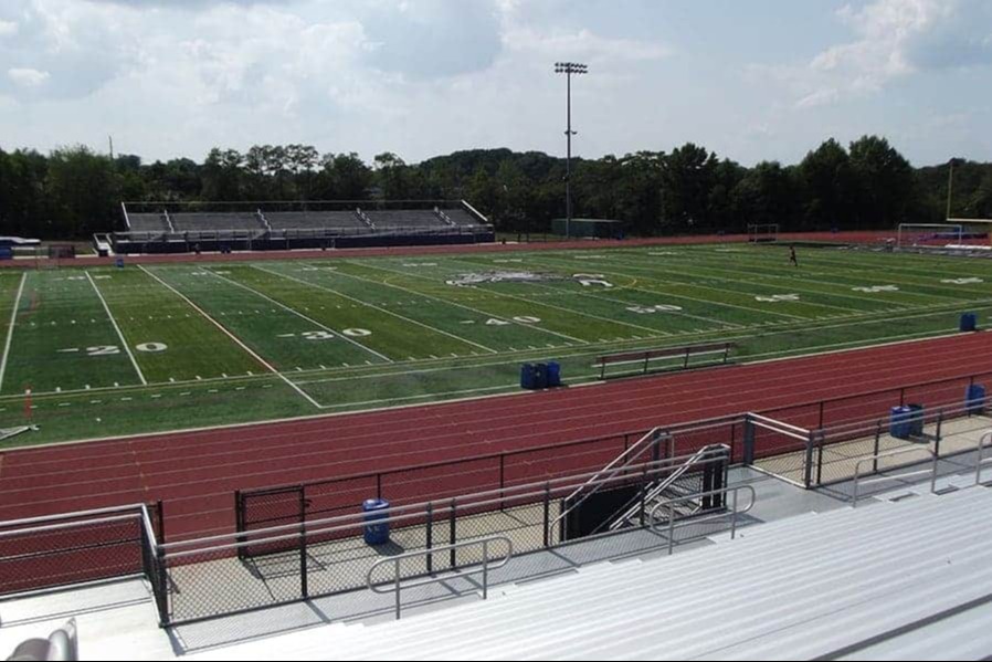 An empty stadium with a football field and track. Green grass covers the field with white markings. Blue barrels are on the track.