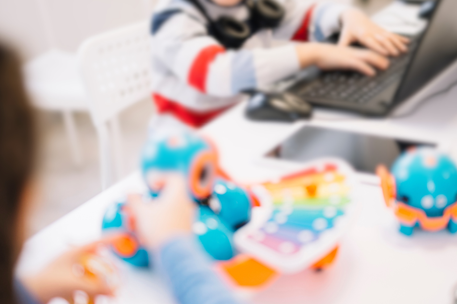 Two children play with toys on a white table, one seated with a laptop and the other standing.