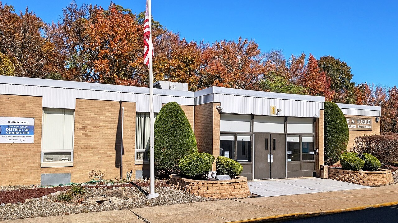 A building with brick walls and a flagpole in front. There are plants and bushes. Trees in various colors are in the background.