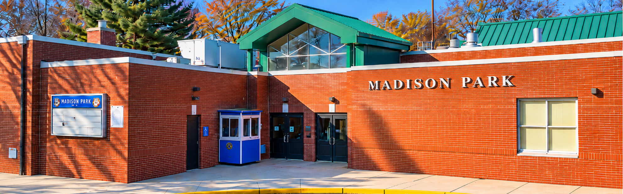 A brick building with green roof, "Madison Park" on top, glass doors, a small booth, and trees in background.