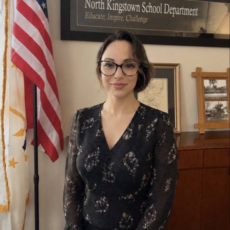 A woman in glasses stands next to a flag and desk. Behind her, a sign reads "North Kingstown School Department."