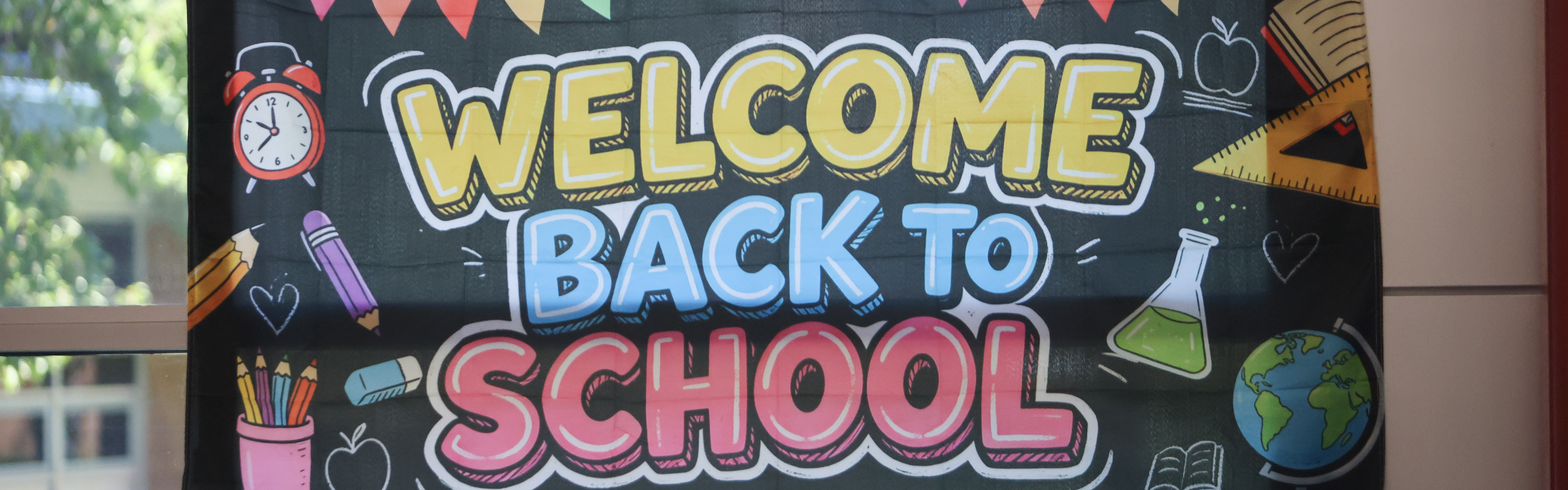 A banner with colorful pencils, a globe, and a clock reads "Welcome Back to School" against a glass window.
