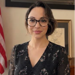 A woman with short hair and glasses, wearing a black floral top, stands in front of a wall with a framed map.