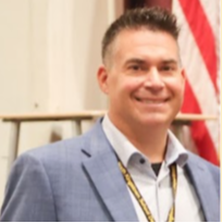 A man in a light blue suit with a white shirt and a yellow lanyard stands smiling. Behind him is an American flag.