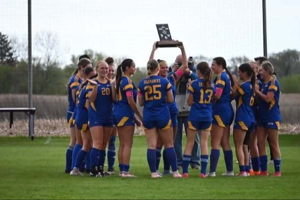 Team in blue uniforms gathers around a trophy on a grassy field with trees and poles in the background.
