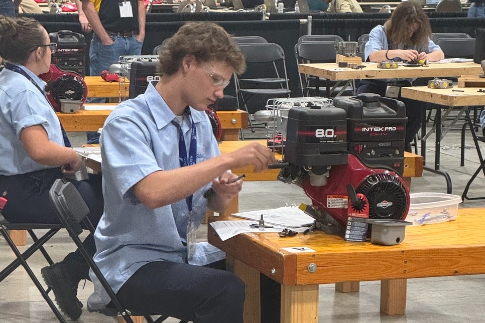 People at a convention with a man seated on a chair at a desk with a machine.