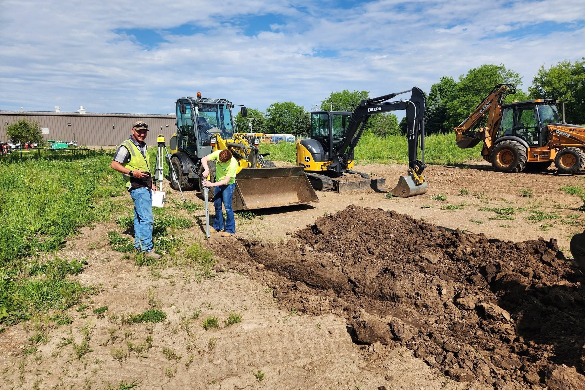 Two individuals in safety vests stand near a dirt pile, with heavy machinery like excavators in background.