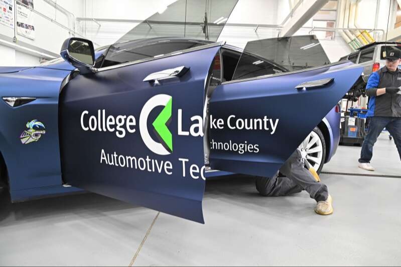 Blue Tesla Model 3 with open door. Logo reads College Lake County Automotive Technologies. A person is crouched next to the car.